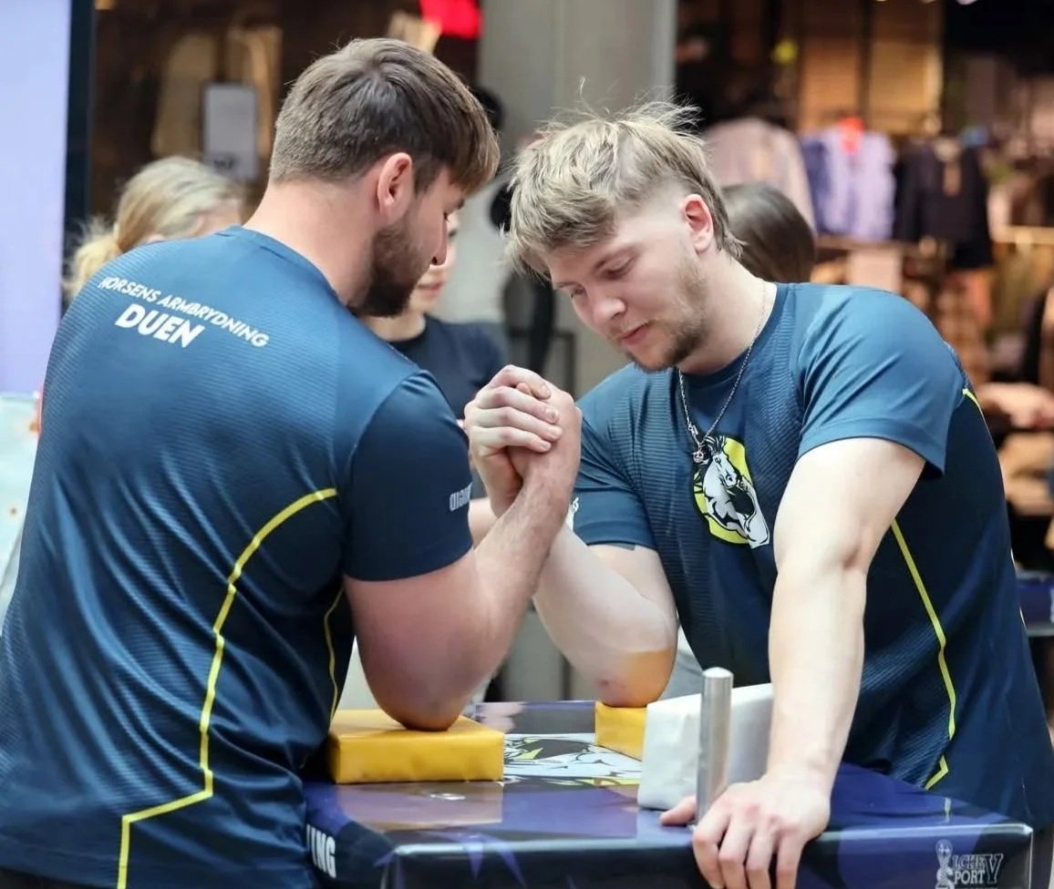 To mænd deltager i arm wrestling konkurrence, iført sportstrøjer med logoer, armene spændt, konkurrerende i et arm wrestling kamp.