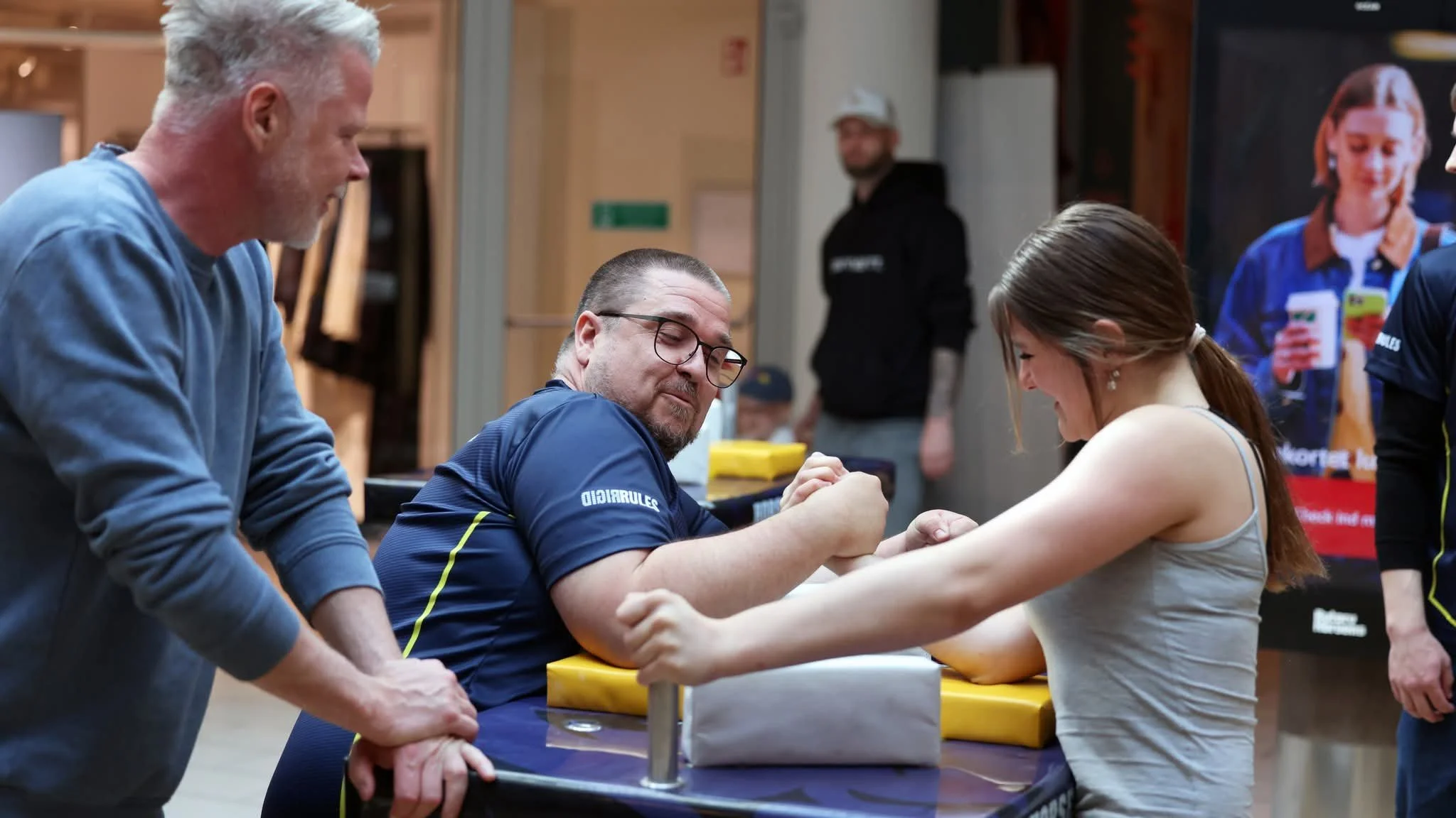 To mænd og en kvinde deltager i arm wrestling konkurrence. To mænd står og holder kvindens hænder, mens hun anstrenger sig for at vinde.