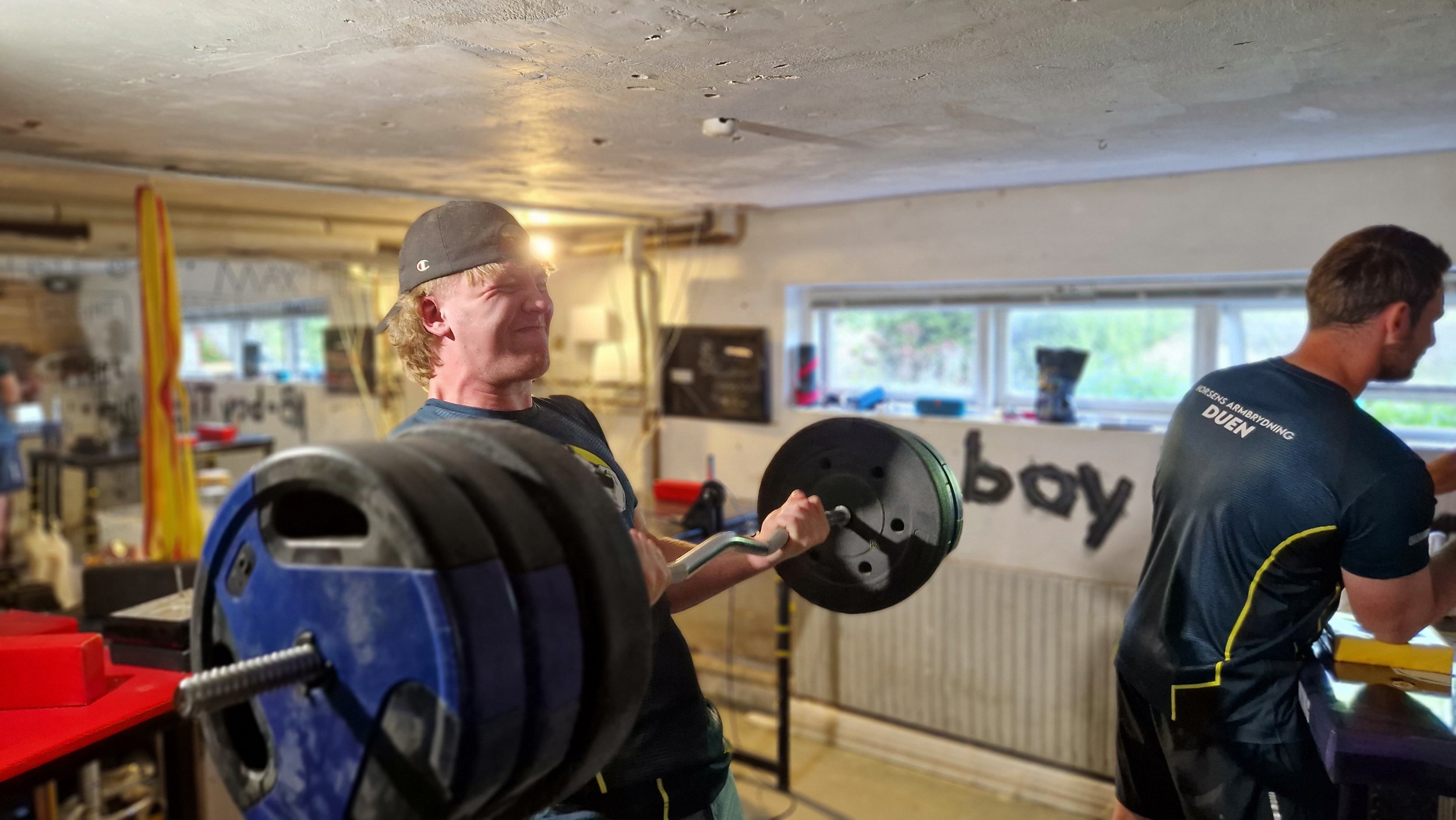 To mænd, der træner med vægte i et fitnesscenter. Den ene løfter en vægtstang, den anden står bag ham.