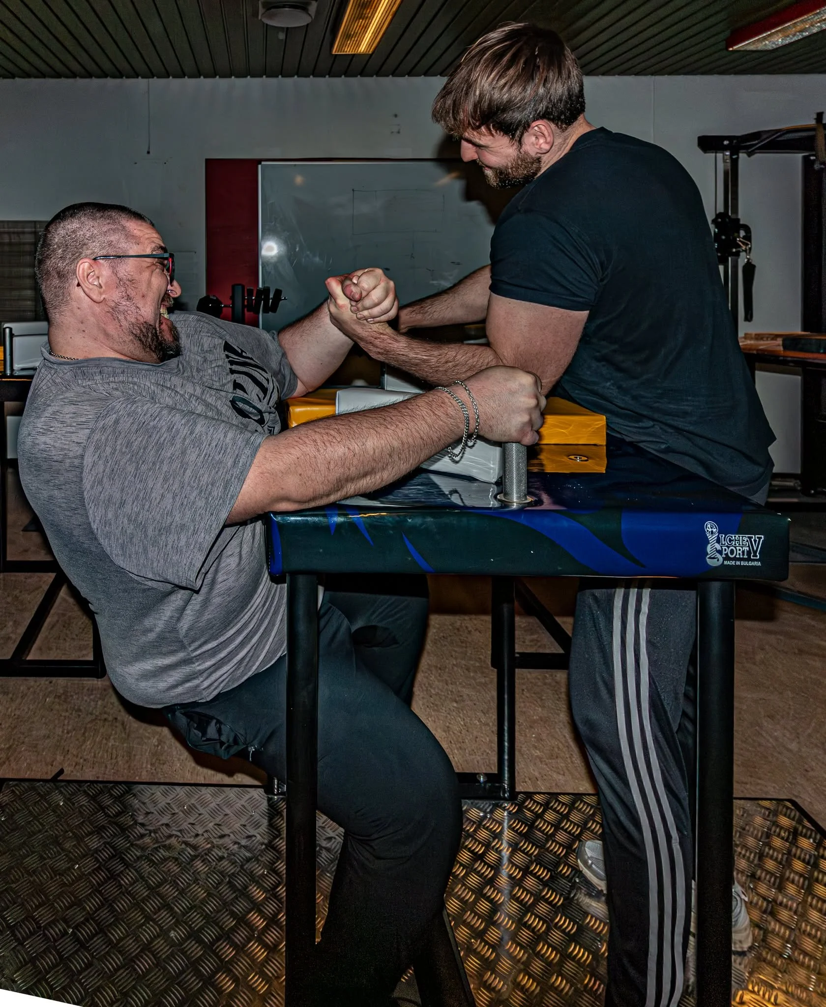 To mænd arm wrestles i et fitnessrum, den ene med skæg, den anden med kort hår, på en sort bord med guldkant.