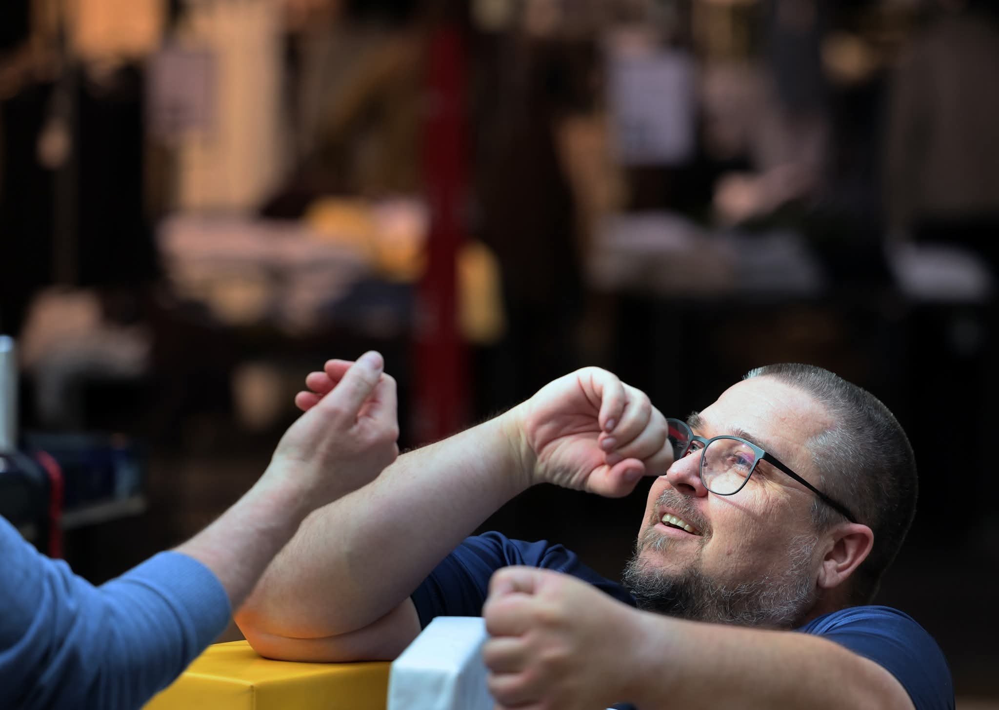 To mænd leger arm wrestling, mens de smiler. De er i et indendørs miljø med en crowd i baggrunden.