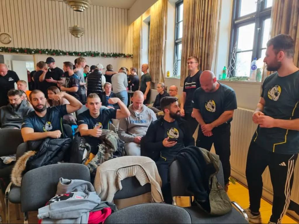 En gruppe mennesker, herunder nogle i matchende t-shirts med en tiger-logo, sidder og står i et rum med store vinduer. Nogle er i samtale, mens andre venter eller slapper af.