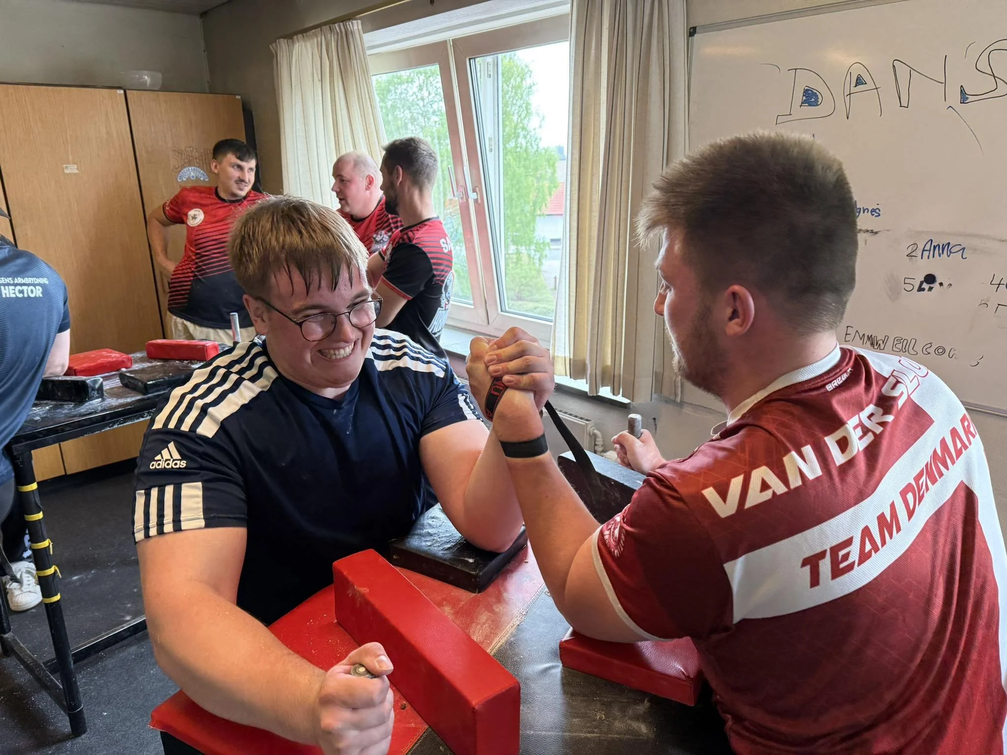 To mænd deltager i arm wrestling konkurrence i et klasseværelse, med flere andre mænd omkring dem, en whiteboard med skrivning i baggrunden og et vindue med grønne træer udenfor.