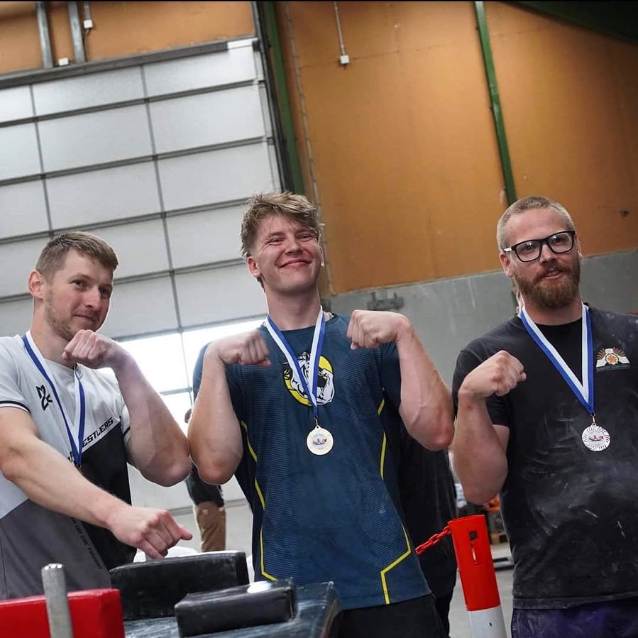 Tre mænd med medaljer omkring halsen, der gør seje muskler med hænderne, står i en industriel eller sportslignende indendørsmiljø.