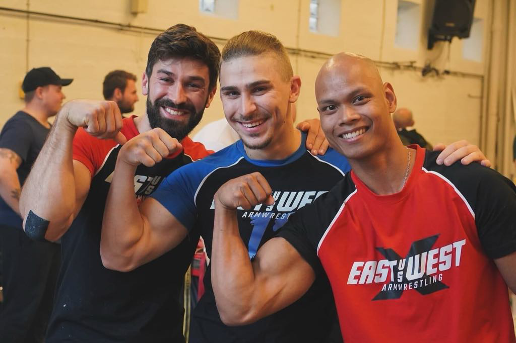 Tre mænd der smiler og poserer flexende deres biceps, iført træningstøj med armwrestling logoer, i et indendørs miljø.