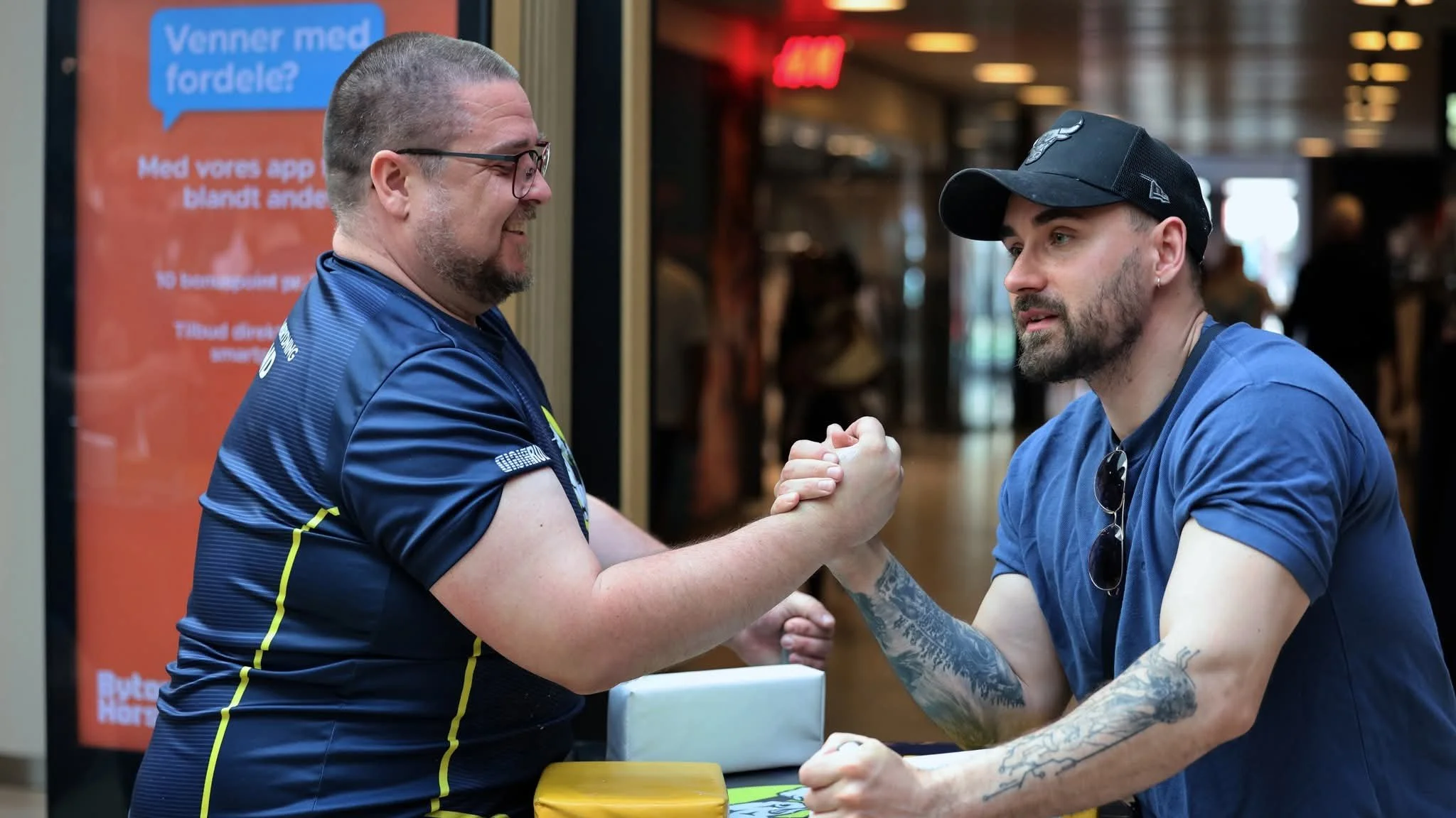 To mænd holder hinanden i hånden i et armrul, mens de smiler. Den ene mand har kort, brunt hår, og den anden har en sort skæg og hætte. De er iført blå T-shirts.