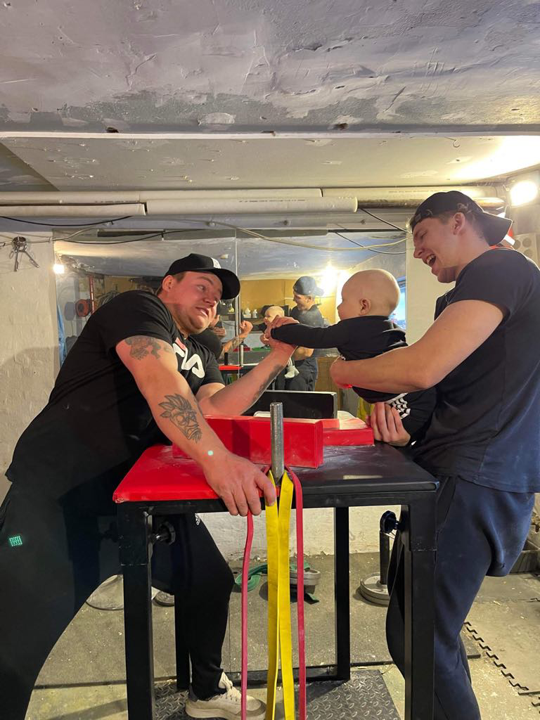 To mænd deltager i arm wrestling i en kælder. En af mændene er barn, som bliver holdt af den anden mand, og de smiler.