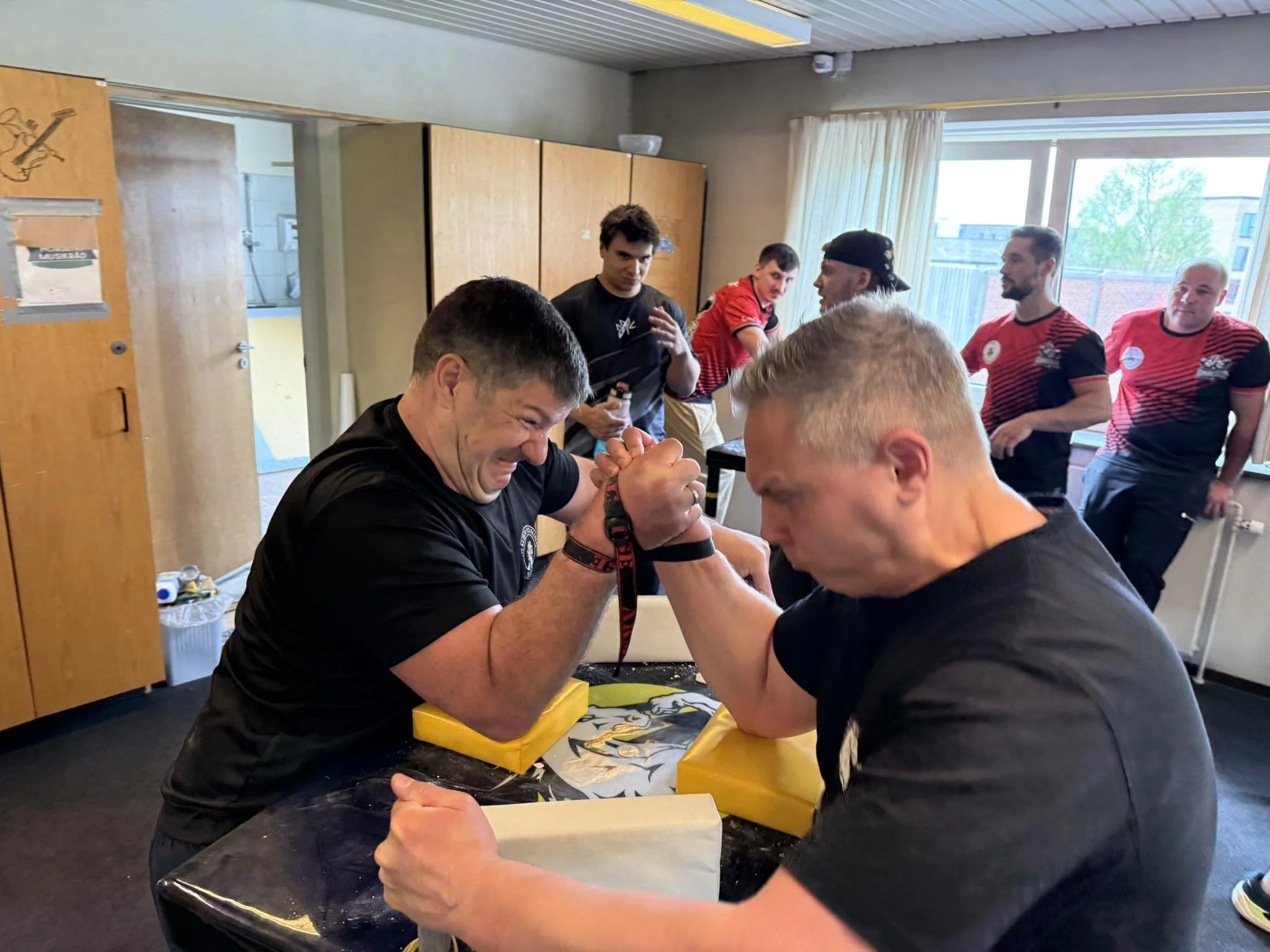 Gruppe personer, der deltager i armwrestling konkurrence i et lyst værelse med store vinduer og gardiner, nogle i sportstøj.