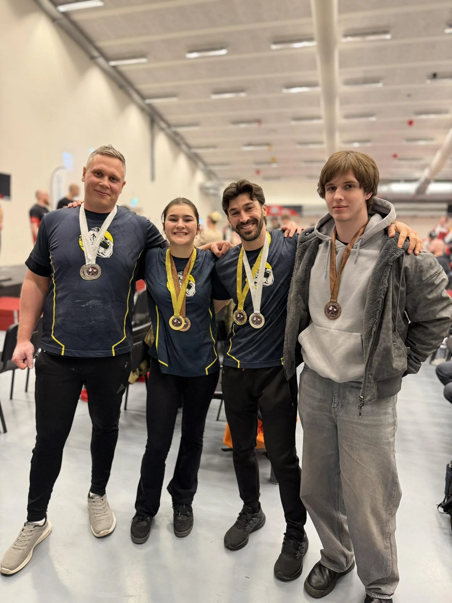 Fire personer står side om side i en indendørs sportsarena og har medaljer rundt halsen, de smiler og har armene på hinandens skuldre.