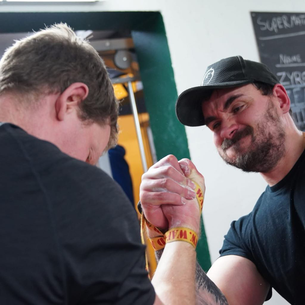 To mænd deltager i arm wrestling konkurrence, hvor den ene mand har på en sort cap og den anden har tykt armbånd og kort hår.