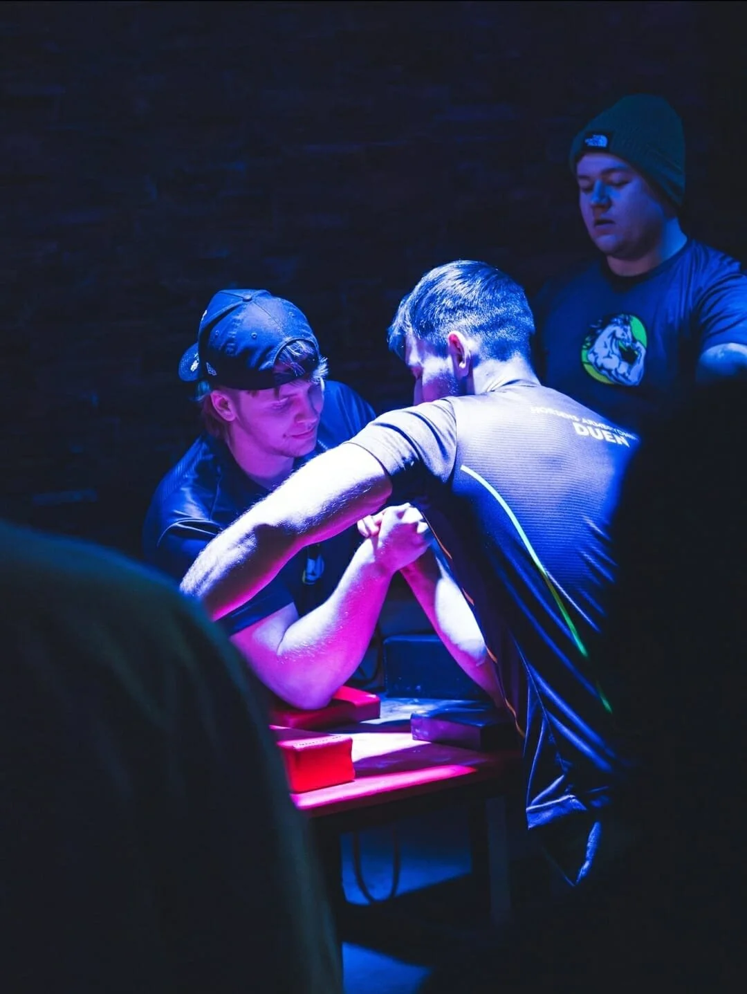 To mænd deltager i arm wrestling under blå belysning, mens en tredje mand ser til.