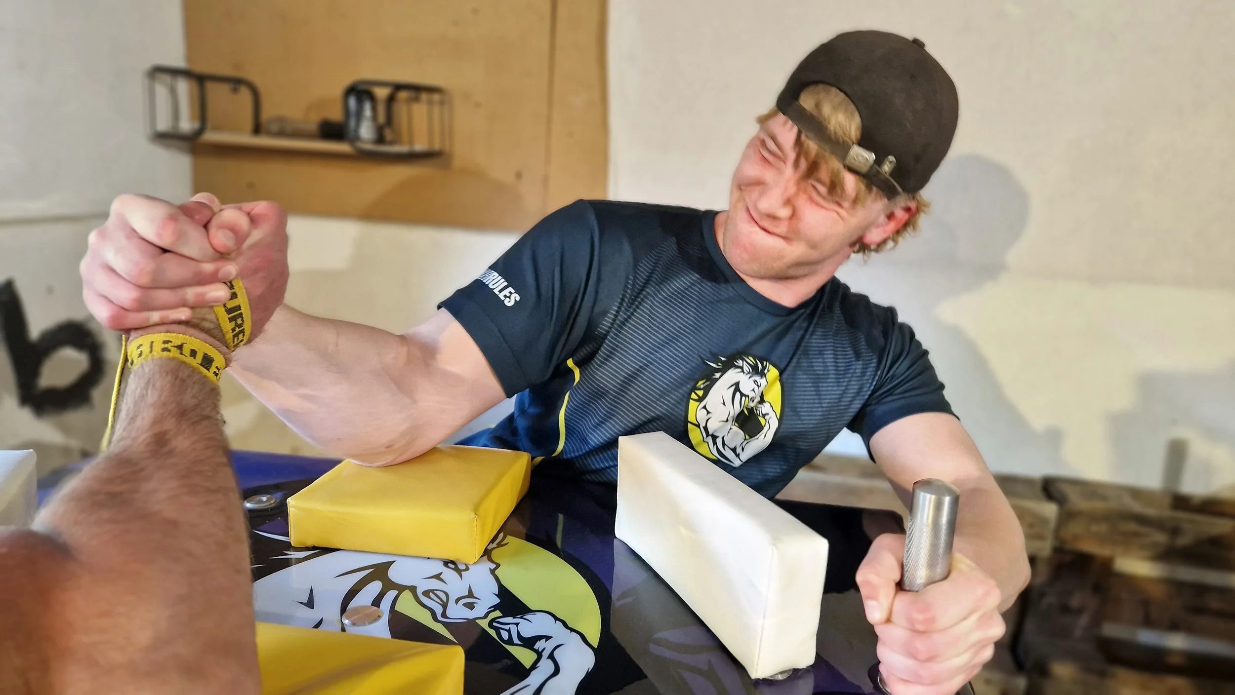 To mænd armwrestles, den ene er yngre med cap og afslappet tøj, de holder hænder i armwrestling, i en indendørs setting.