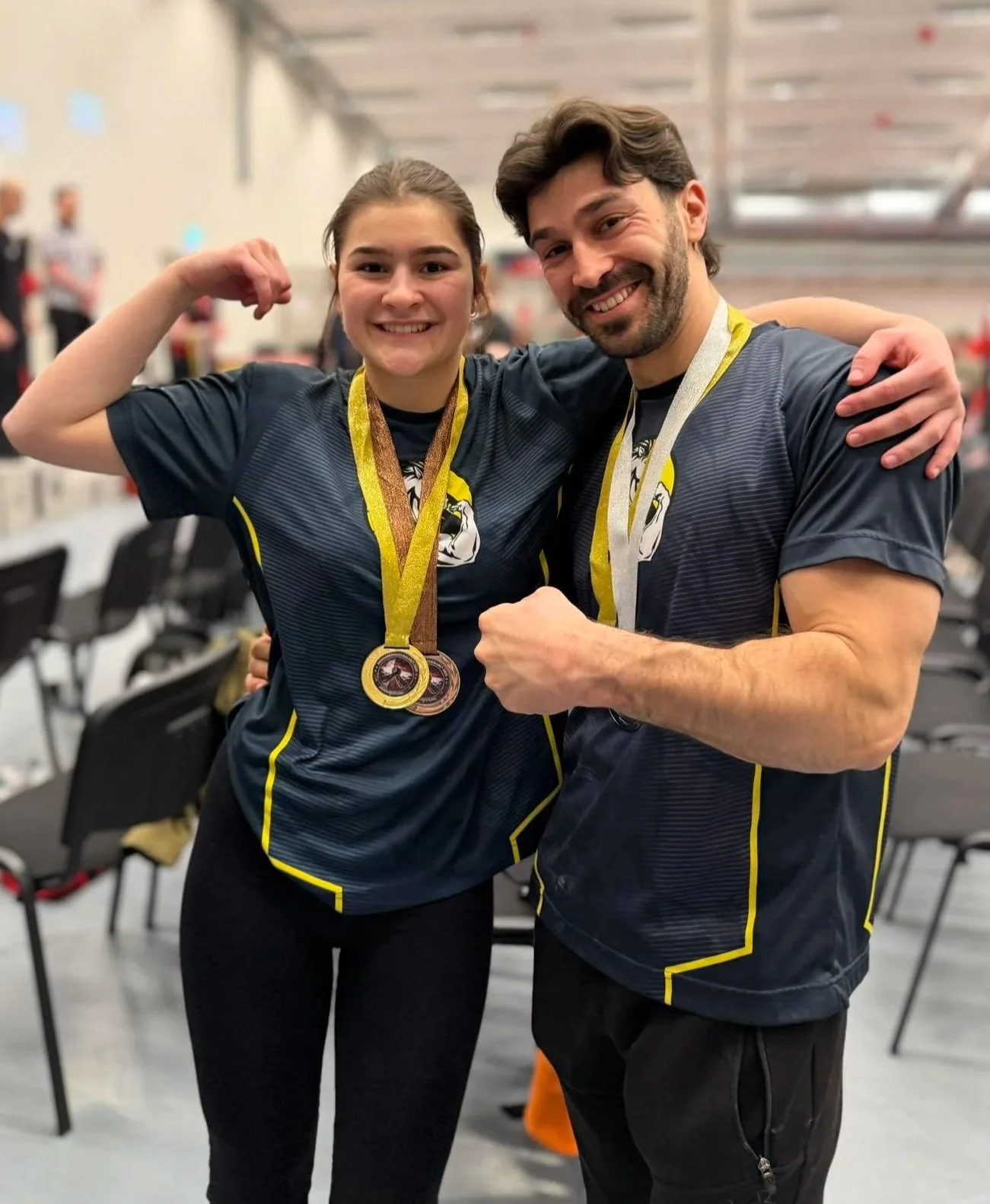 To mennesker iført sportstøj foran en baggrund med stole, de viser deres medaljer og har armene omkring hinanden i en triumferende positur.