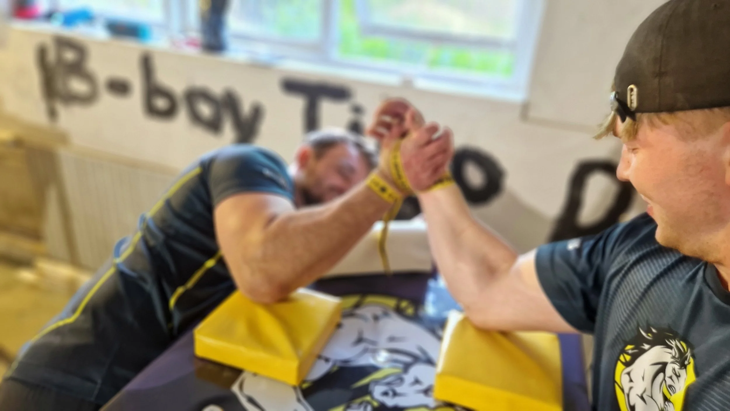 To mænd deltager i arm wrestling konkurrence i et rum med vindue i baggrunden, hvor en af mændene smiler. De er klædt i sportsatletik og sidder ved et bord.