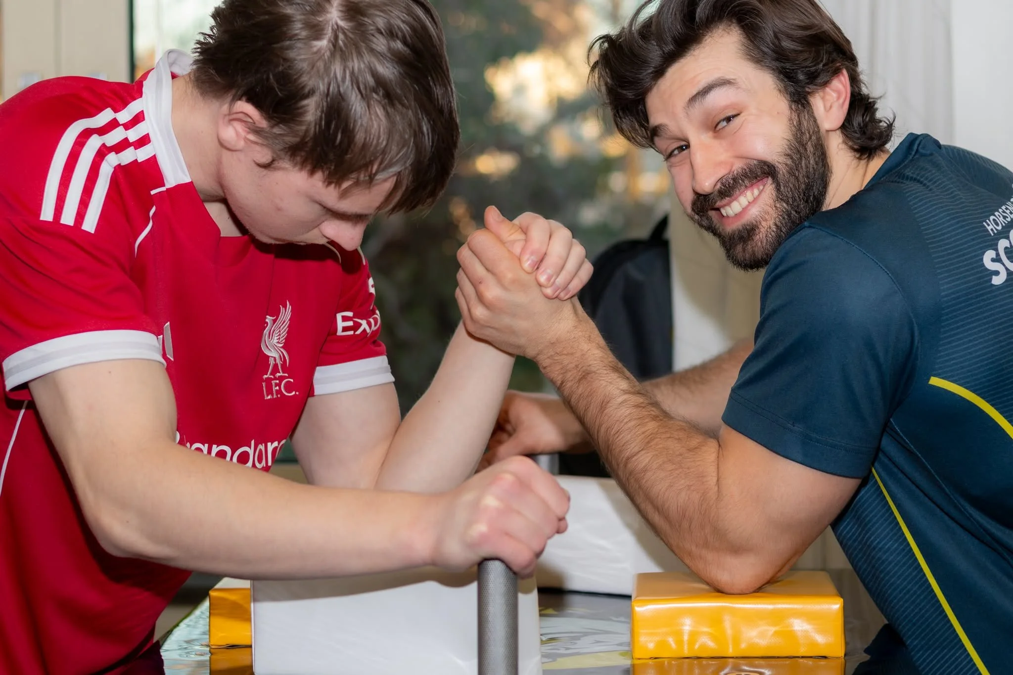 To mænd deltager i arm wrestling konkurrence, den ene i rød hjemmehold trøje, den anden i blå uniform, i et harmonisk og smilende øjeblik.