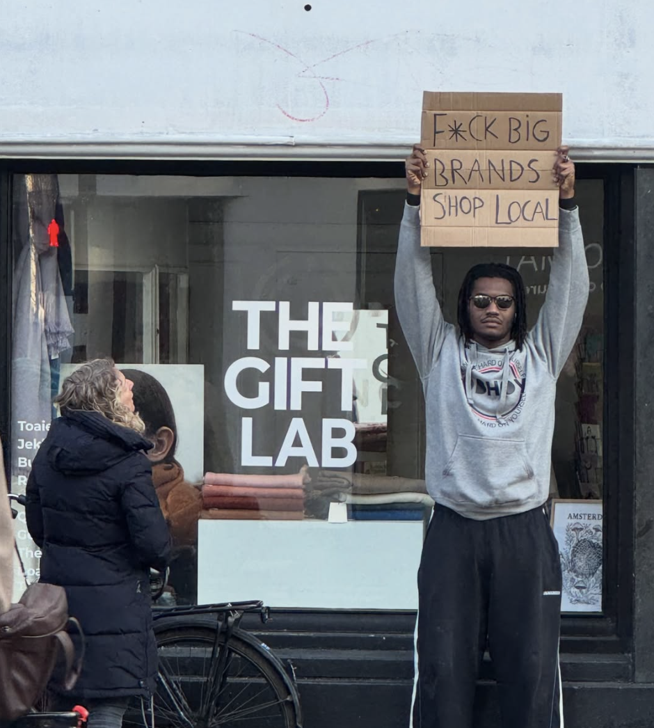 Man met dreadlocks en zonnebril houdt een bord omhoog met de tekst 'F*CK BIG BRANDS SHOP LOCAL' voor een winkel met de naam 'THE GIFT LAB'.