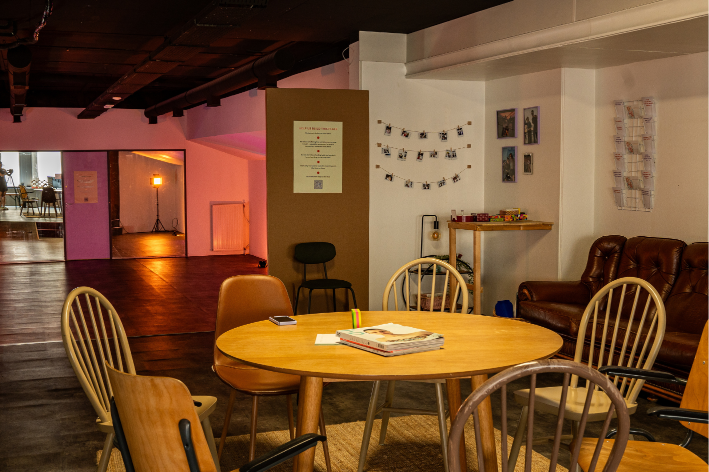 Interieur van een gezellige kamer met een ronde houten tafel, omringd door verschillende stoelen en banken, decoraties aan de muur, en een open ruimte met een lamp en een deur in de achtergrond.