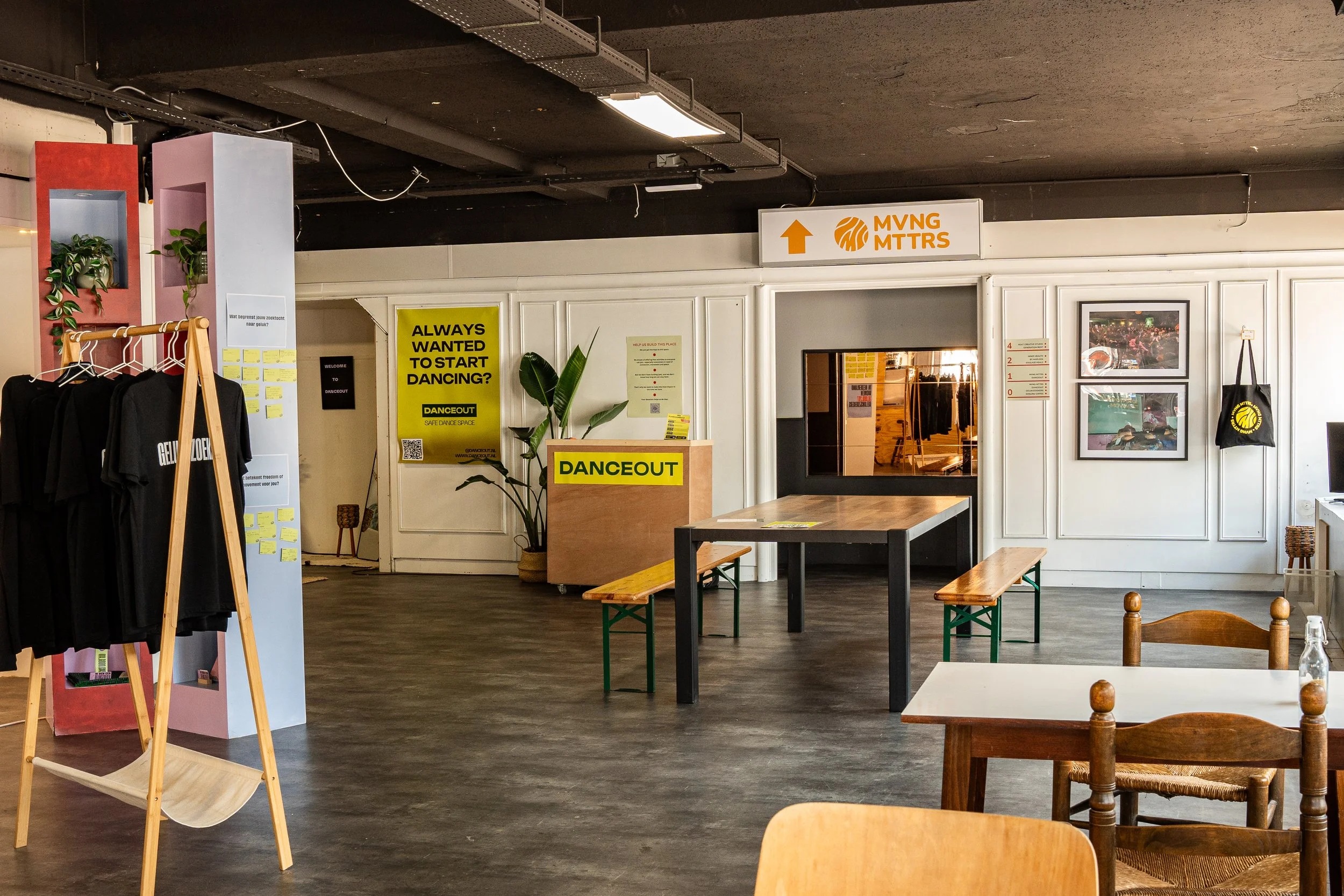 Binnenruimte met tafels, stoelen, planten, en winkelinrichting. Signalen en decoraties hangen aan de muur, en er is een deur met ramen.