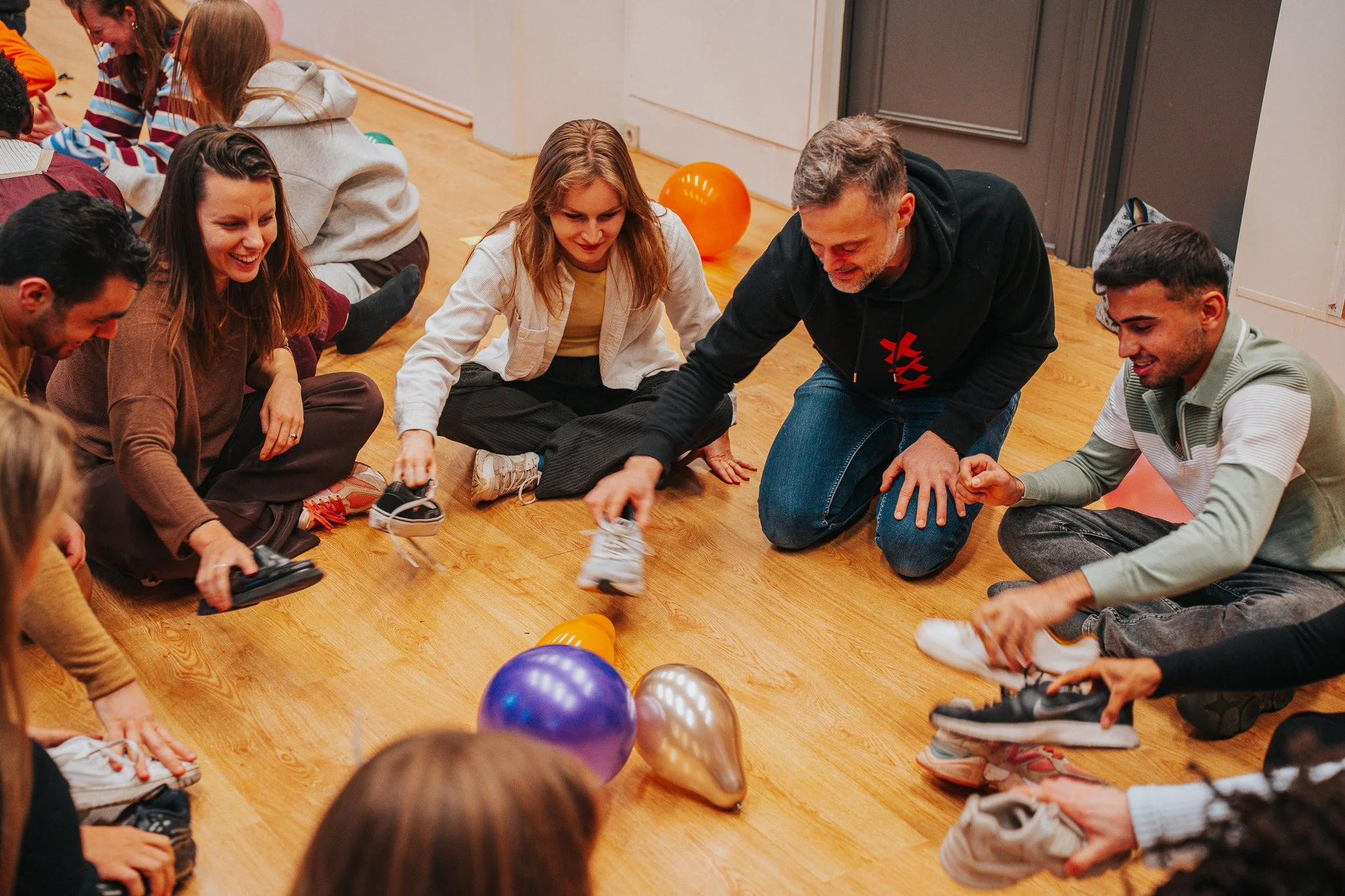 Groep mensen zitten in een cirkel op de vloer, lachen en spelen met ballonnen tijdens een feestje of bijeenkomst.