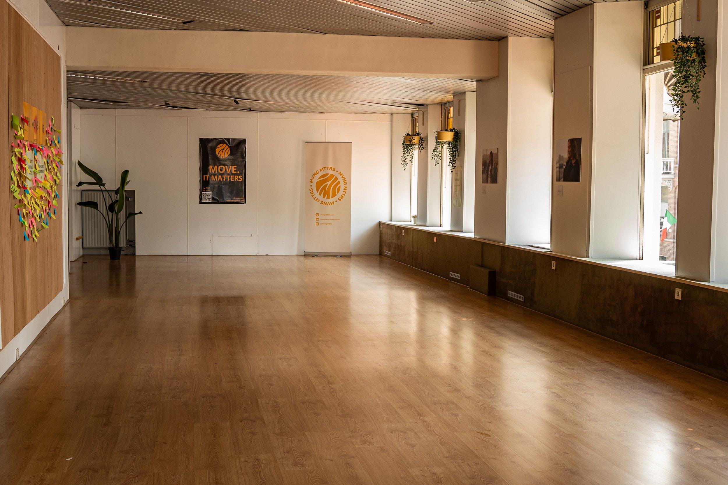 Een lege kamer met houten vloer, witte muren en grote ramen met planten op vensterbanken.
