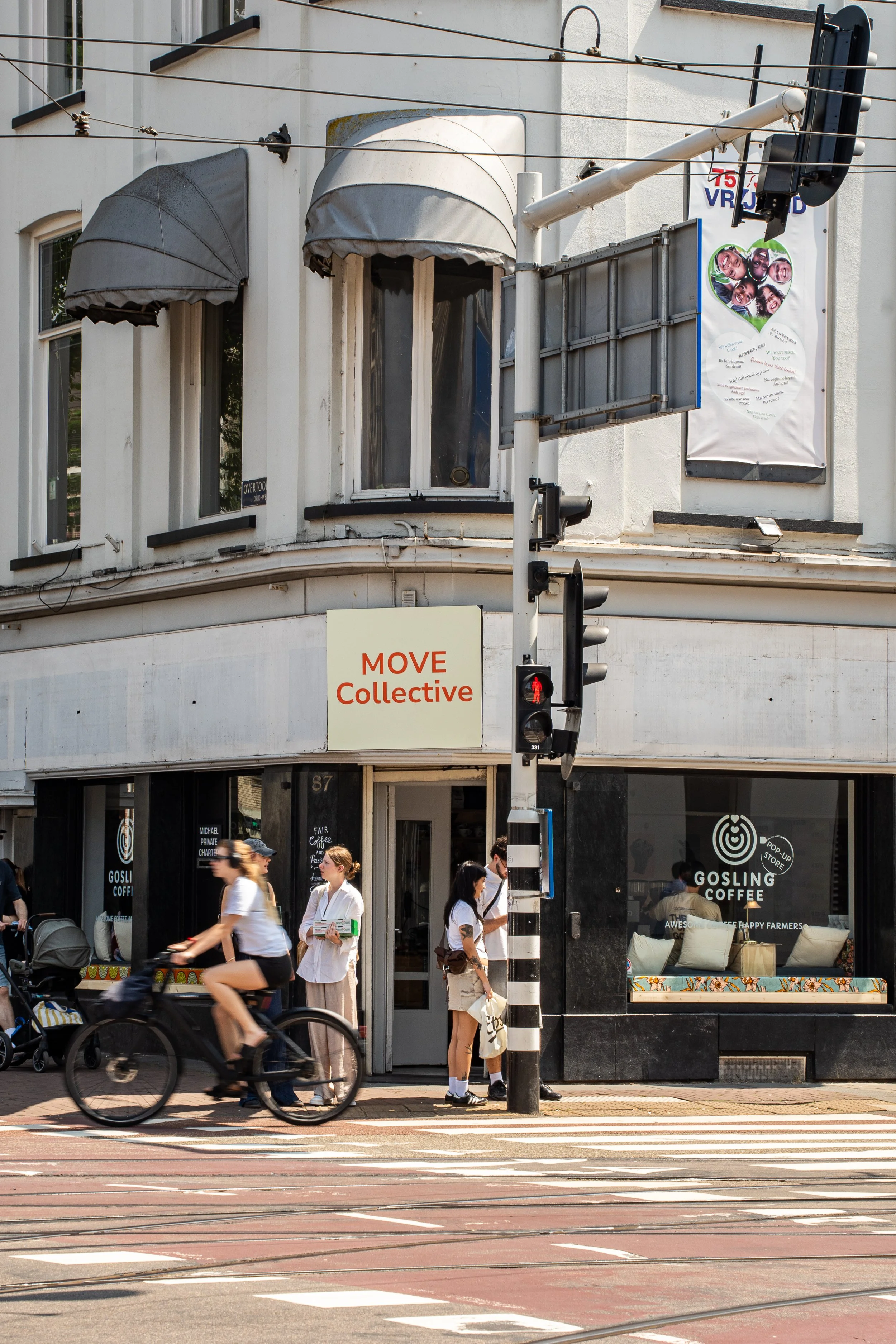 Stadsscene met mensen die wachten bij een oversteek, een persoon fietst voorbij, een koffiebar genaamd Gosling Coffee op de achtergrond, een straatverkeerslicht en een gebouw met ramen en posters.