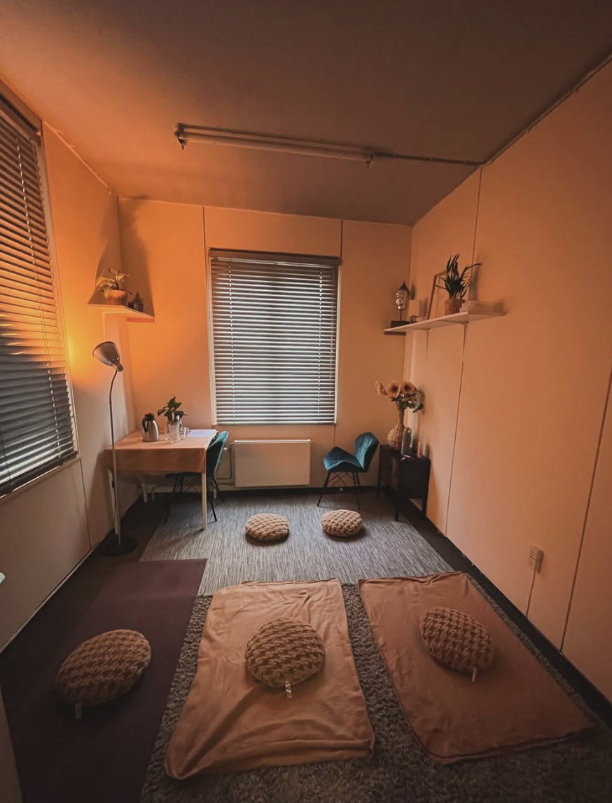 Een rustige meditatiekamer met tapijten en zitzakken, een tafel met bloemen, een boekenplank, en raambedekking met horizontale jaloezieën.