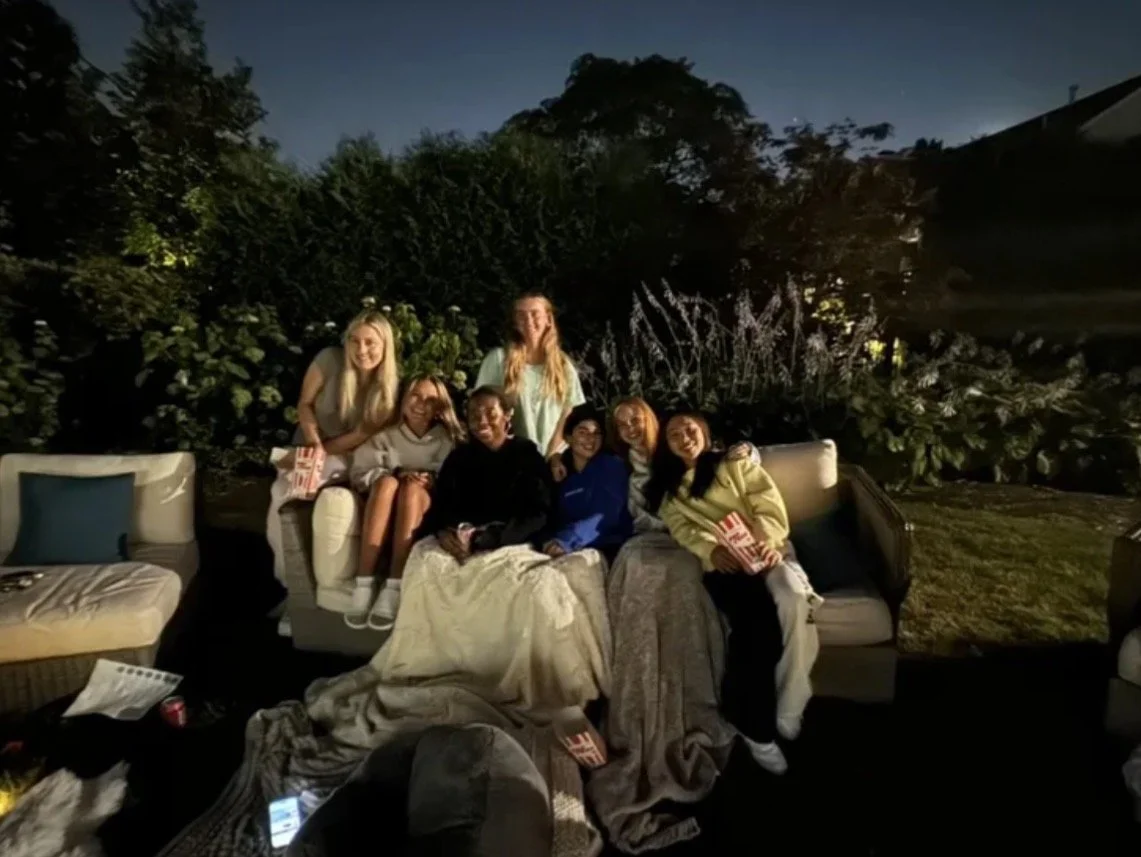 Group of seven women sitting and standing together outdoors on a couch at night, smiling, with trees and bushes in the background.