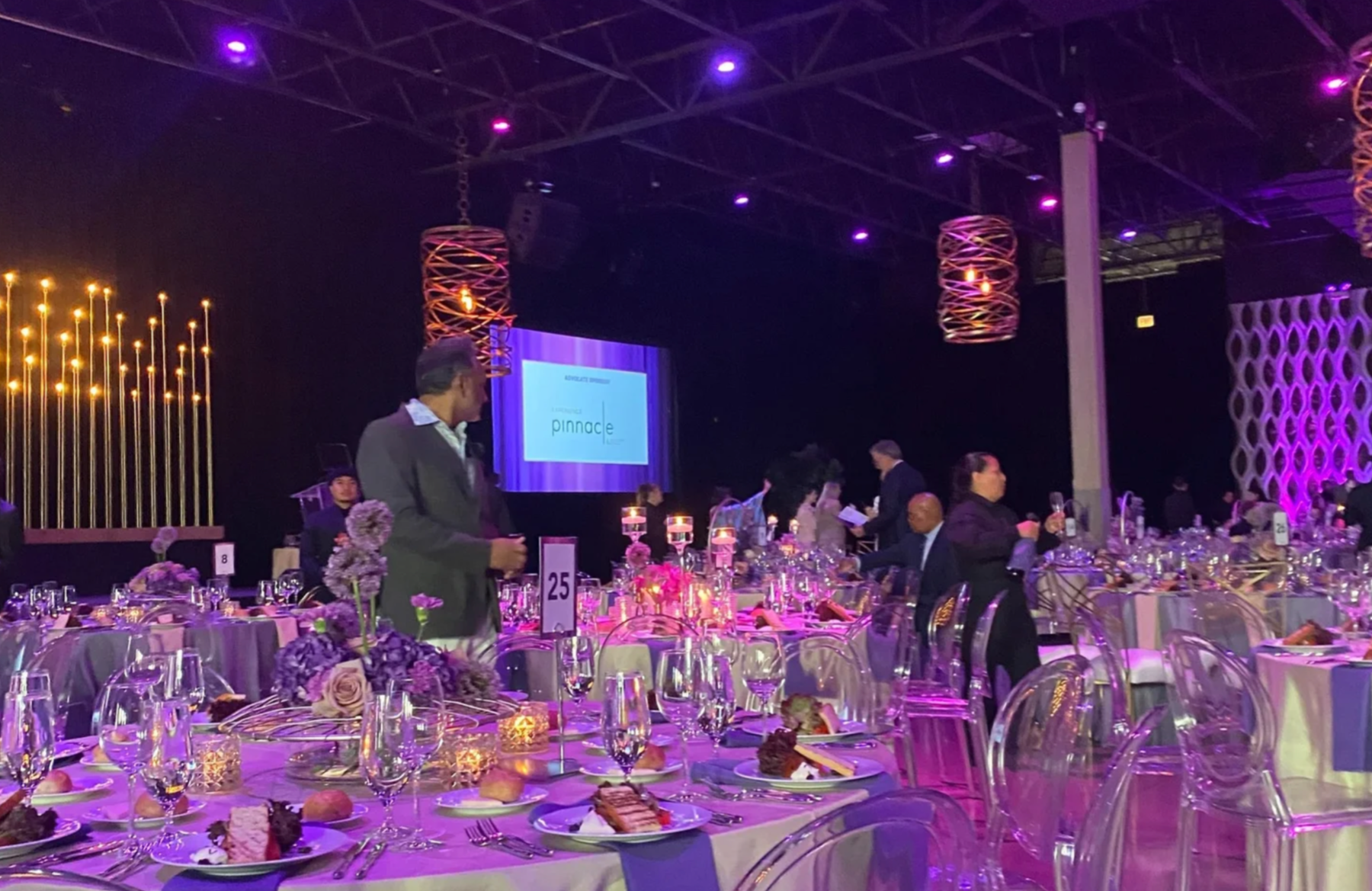 A banquet hall with round tables set for an event, decorated with purple flowers, candles, and purple lighting. People are mingling, and a large screen displays the word 'pinnacle' in the background.