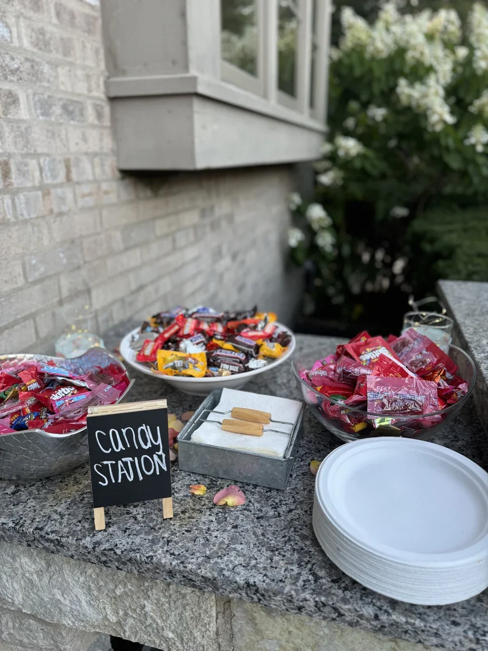 A candy station set up outdoors on a stone table with bowls of various candies, a small chalkboard sign, and a stack of white plates, with a background of a brick wall, window, and greenery.