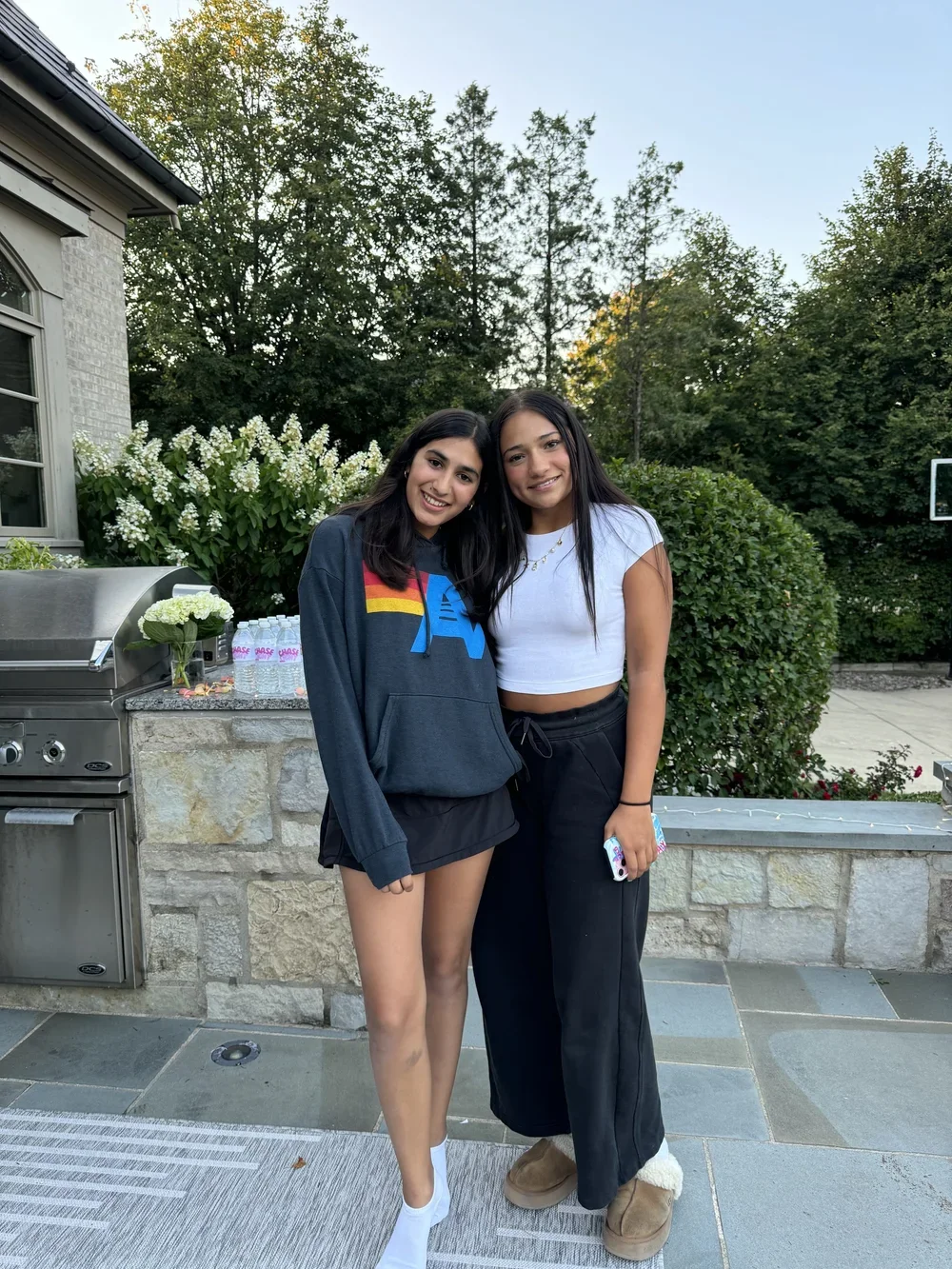 Two young women standing outdoors, smiling, with trees and bushes in the background. One is wearing a dark hoodie with a colorful design, black shorts, and white shoes. The other is wearing a white crop top, black pants, and tan slippers, holding a phone.