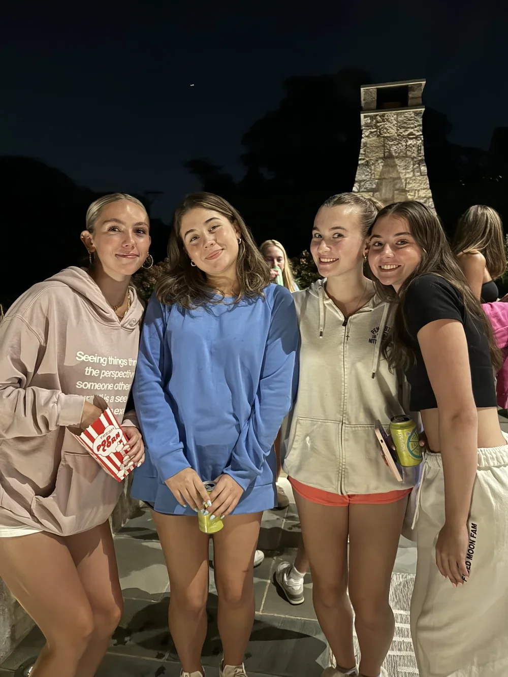 Four young women smiling at night outdoors, with a stone tower in the background, holding drinks and snacks.