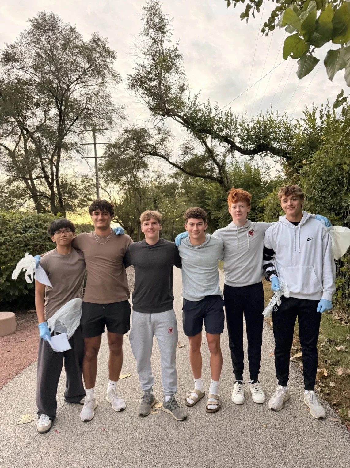 Six young men standing outdoors on a cloudy day, with trees and bushes in the background. They have their arms around each other's shoulders, smiling at the camera. They are wearing casual activewear, some with gloves, and appear to have been involved in a cleanup or volunteer activity.