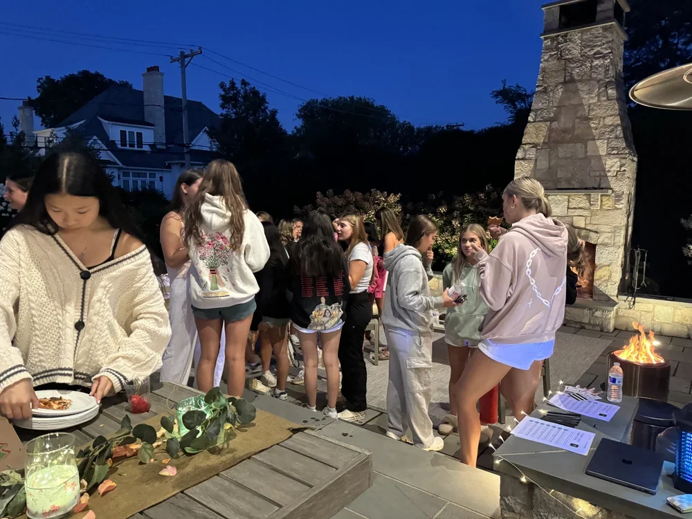 A group of young women gathered outdoors near a stone fireplace at dusk, some conversing, others serving or waiting in line, with a table of food and drinks nearby.
