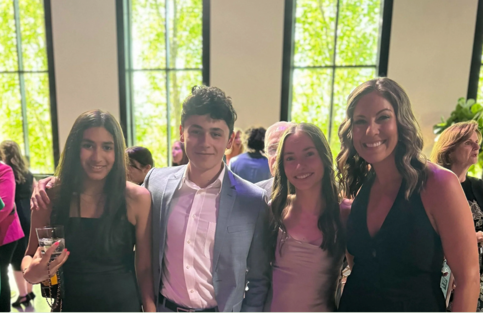 Group of four young adults standing together at an indoor event, smiling, dressed semi-formally, with large windows showing greenery in the background.