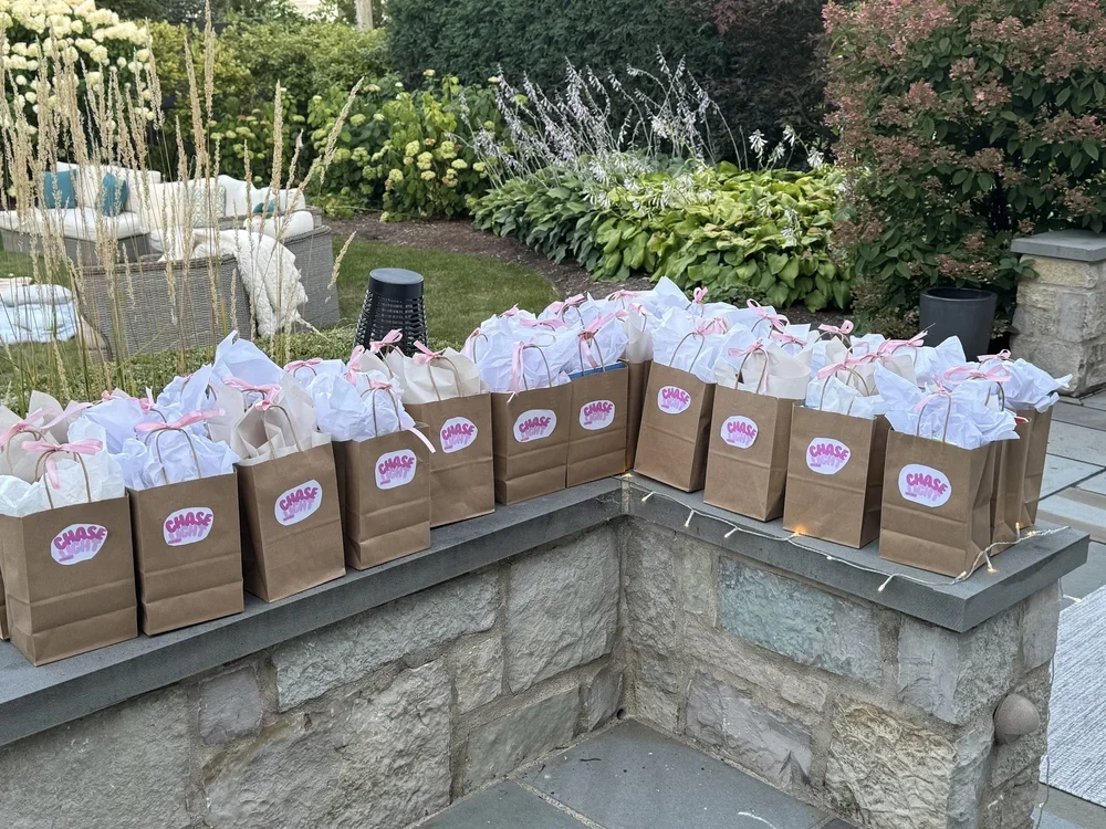 Multiple paper gift bags with pink ribbons and Chase branding, topped with white tissue paper, arranged on a stone ledge in a garden.