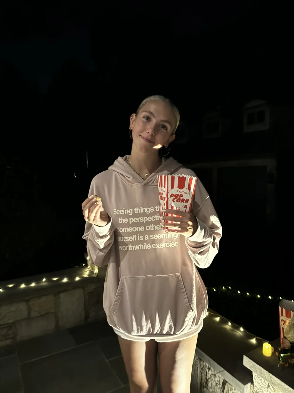 A young woman with short blonde hair holding a large tub of popcorn and a small snack, standing outdoors at night with decorative string lights behind her.