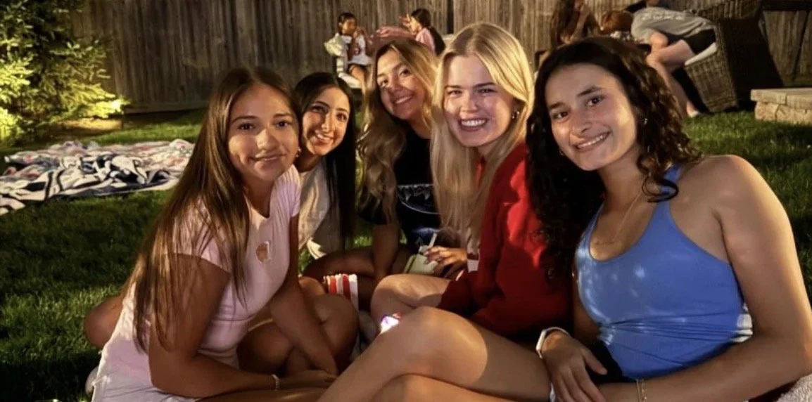Six young women sitting outdoors on the grass, smiling at the camera.