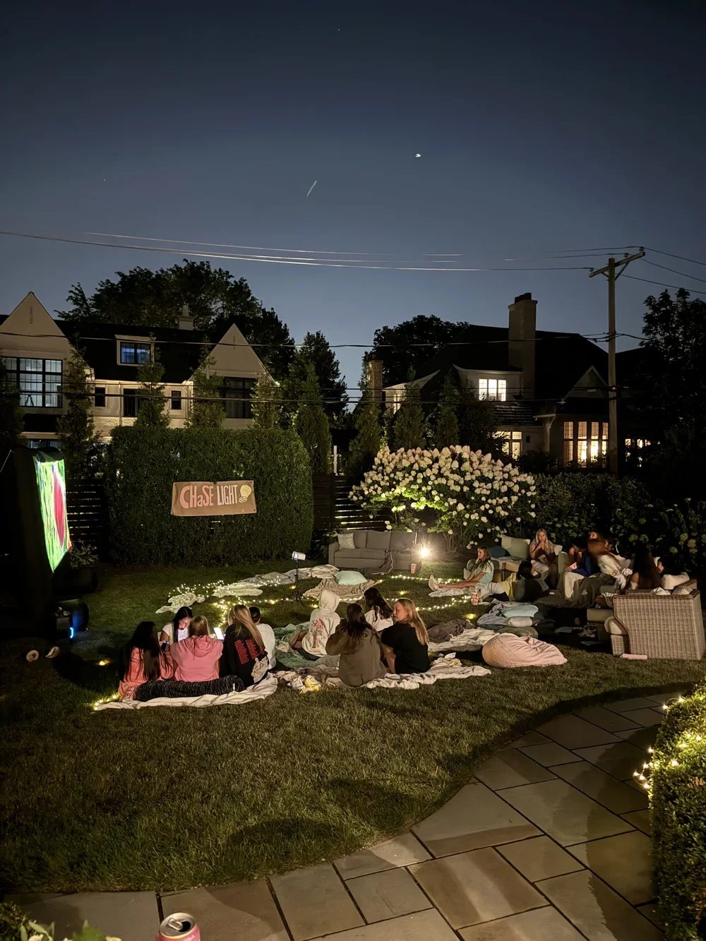 People gathered in a backyard at night for a movie night, sitting on blankets and pillows, with string lights, a large screen, and a banner that reads 'Chase Light'.