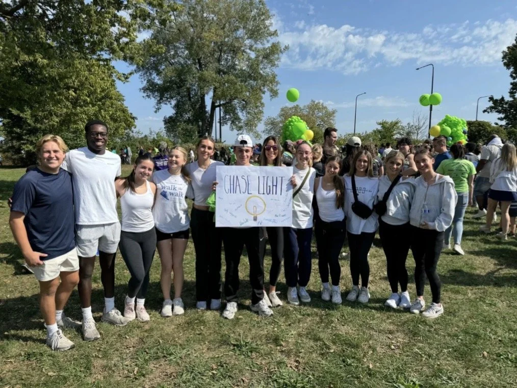 Group of people outdoors, some holding a sign that says 'Chase Light', with green balloons and trees in the background, bright sky.