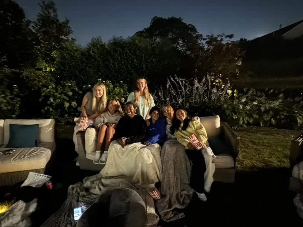 Group of seven women sitting and standing on outdoor couches at night, surrounded by trees and bushes, enjoying a gathering.