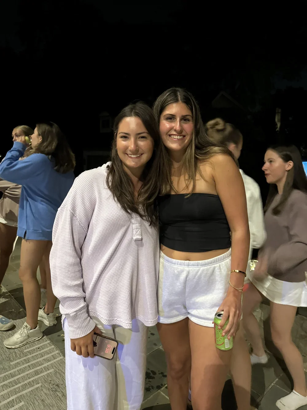 Two young women standing together at night, smiling, with other people in the background.