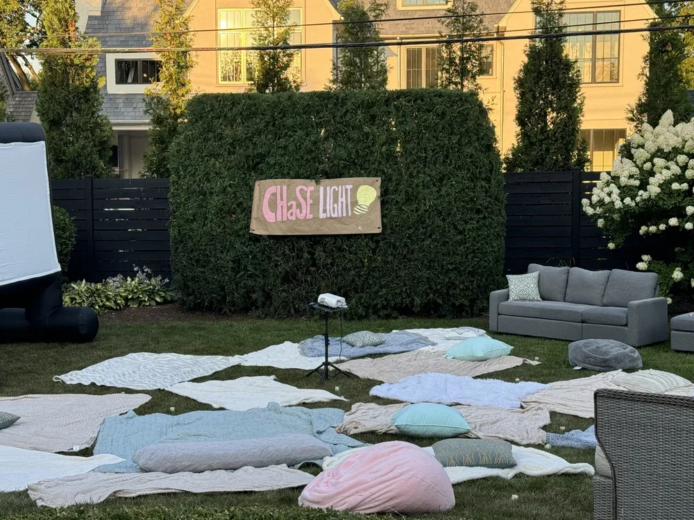 Outdoor shot of a backyard with a makeshift outdoor movie setup called 'Chase Light'. There are blankets and pillows spread on the grass, a projector on a small stand, and a banner with the event name. A gray sofa and a white and gray chair are also present, along with lush green trees and a black fence in the background.