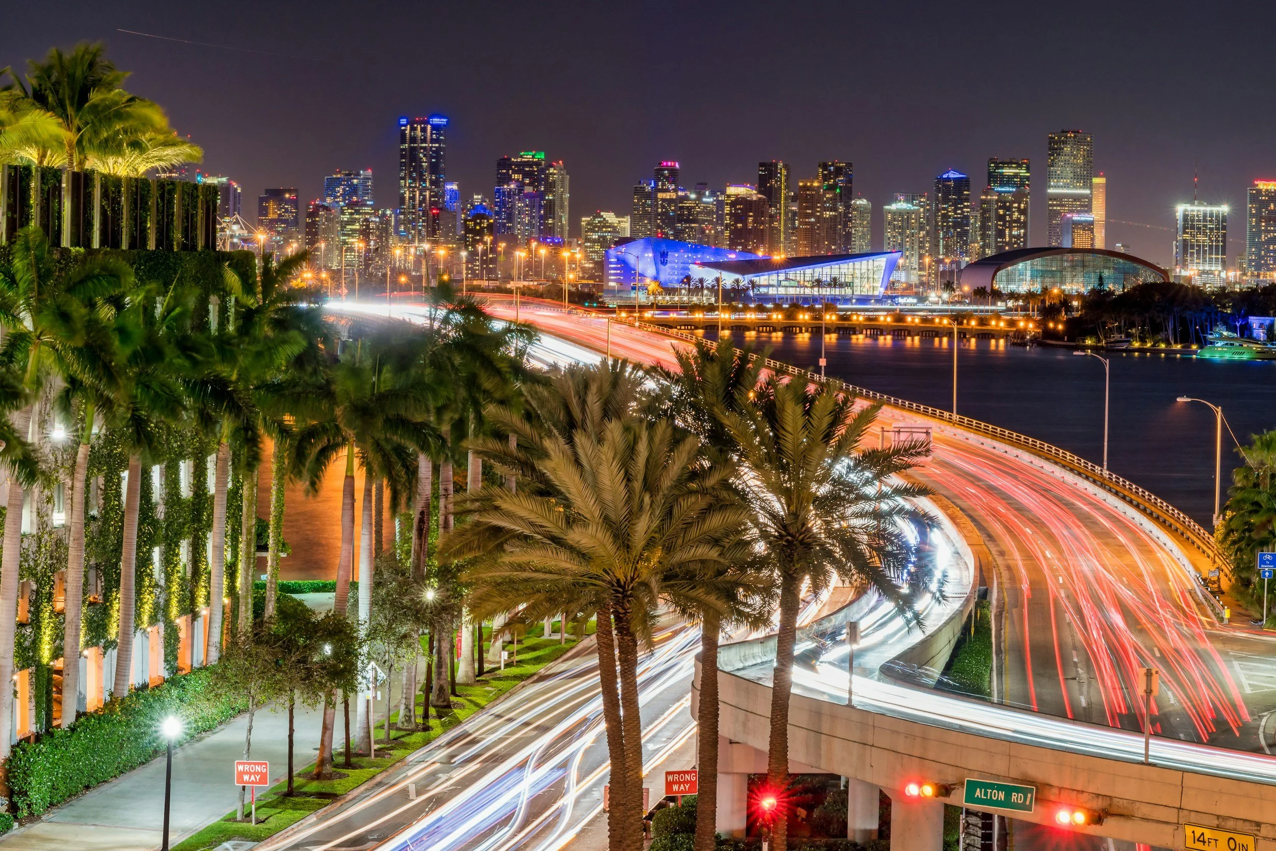 Nighttime cityscape of a downtown with illuminated skyscrapers, palm trees, and long-exposure light trails from traffic on an overpass, with signs indicating 'Wrong Way' and street name 'Alton Rd'.