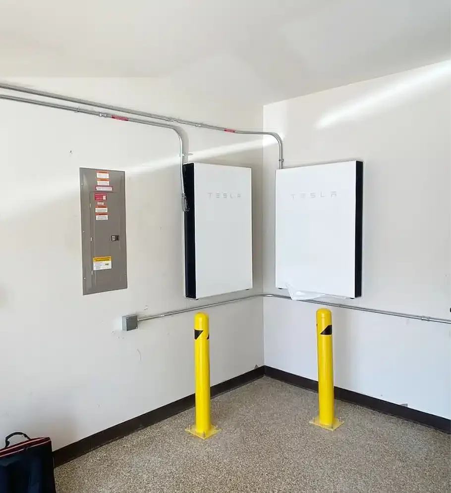 Two Tesla Powerwall batteries mounted on a white wall in a utility room, with a gray electrical panel and yellow protective posts in front.