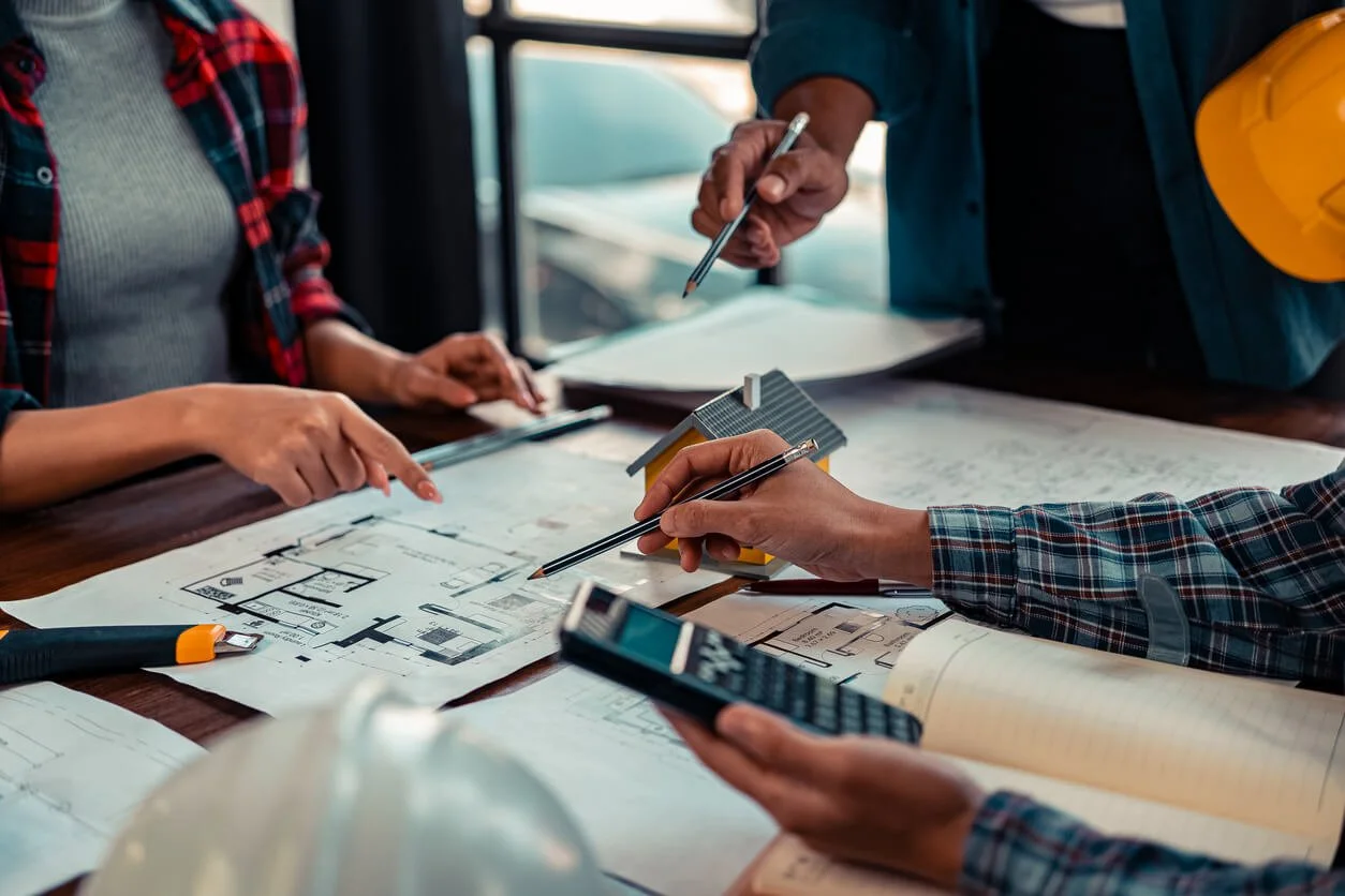 People reviewing architectural plans, with small house model and a calculator on a table.