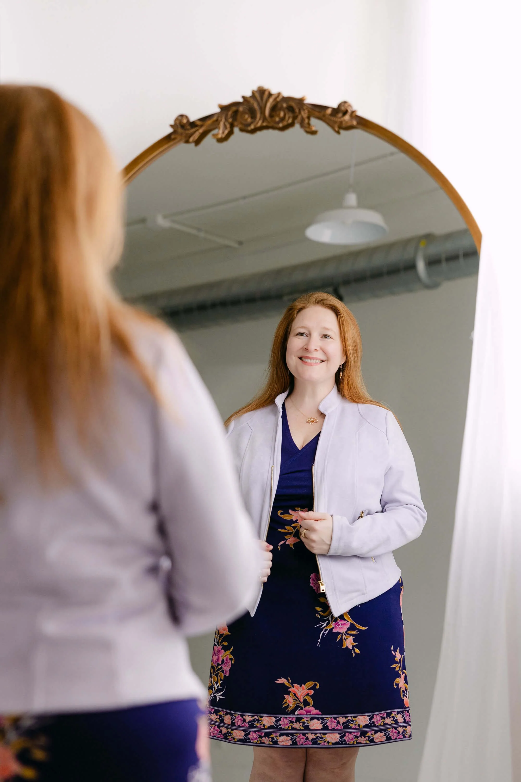 A woman with red hair is smiling while looking at her reflection in a mirror. She is wearing a white jacket over a dark dress with floral patterns, and her hands are clasped in front of her.
