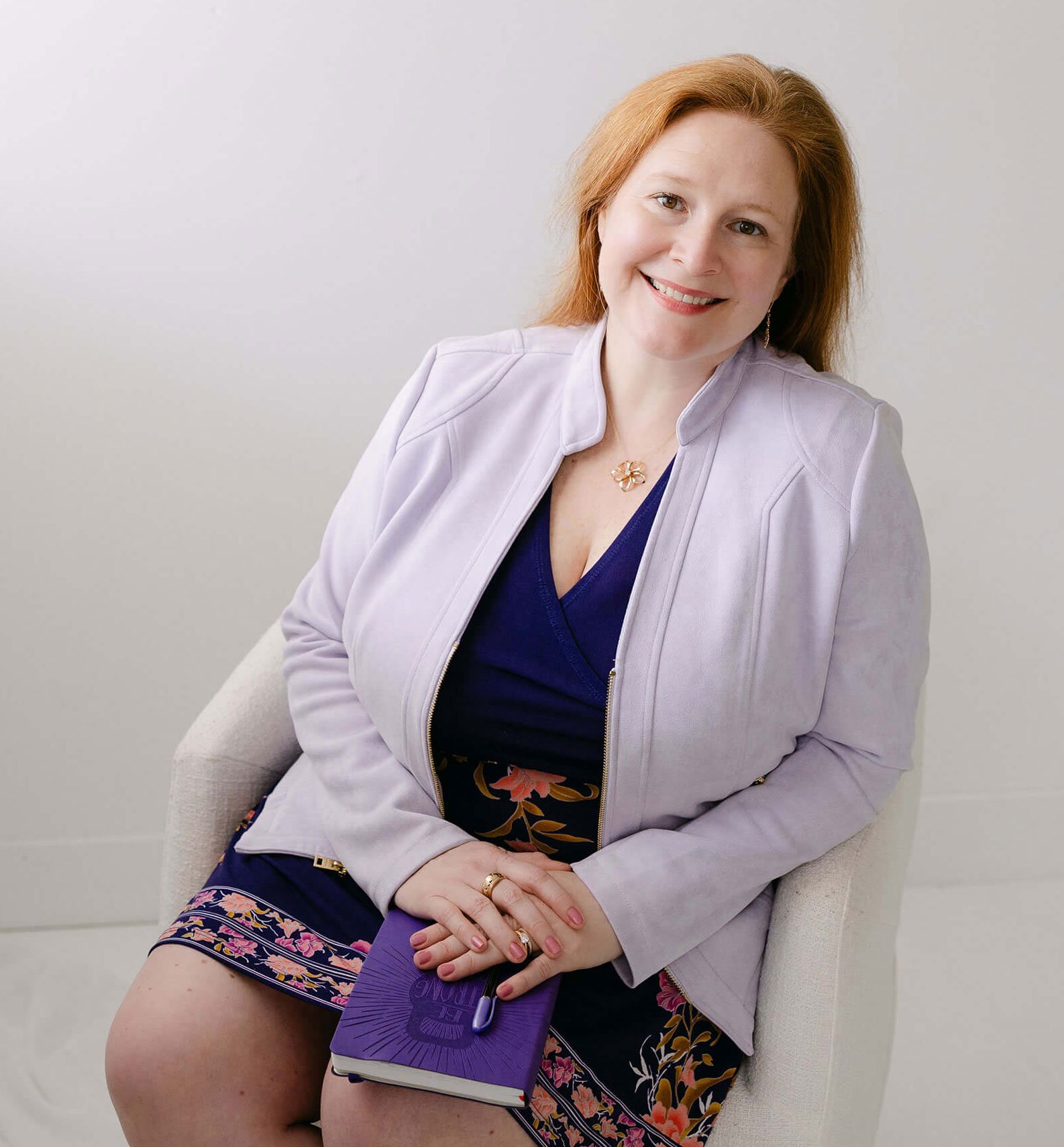 A woman with red hair sitting in a beige armchair, holding a purple fan, wearing a dark blue dress, a light gray jacket, and floral shorts, smiling at the camera.