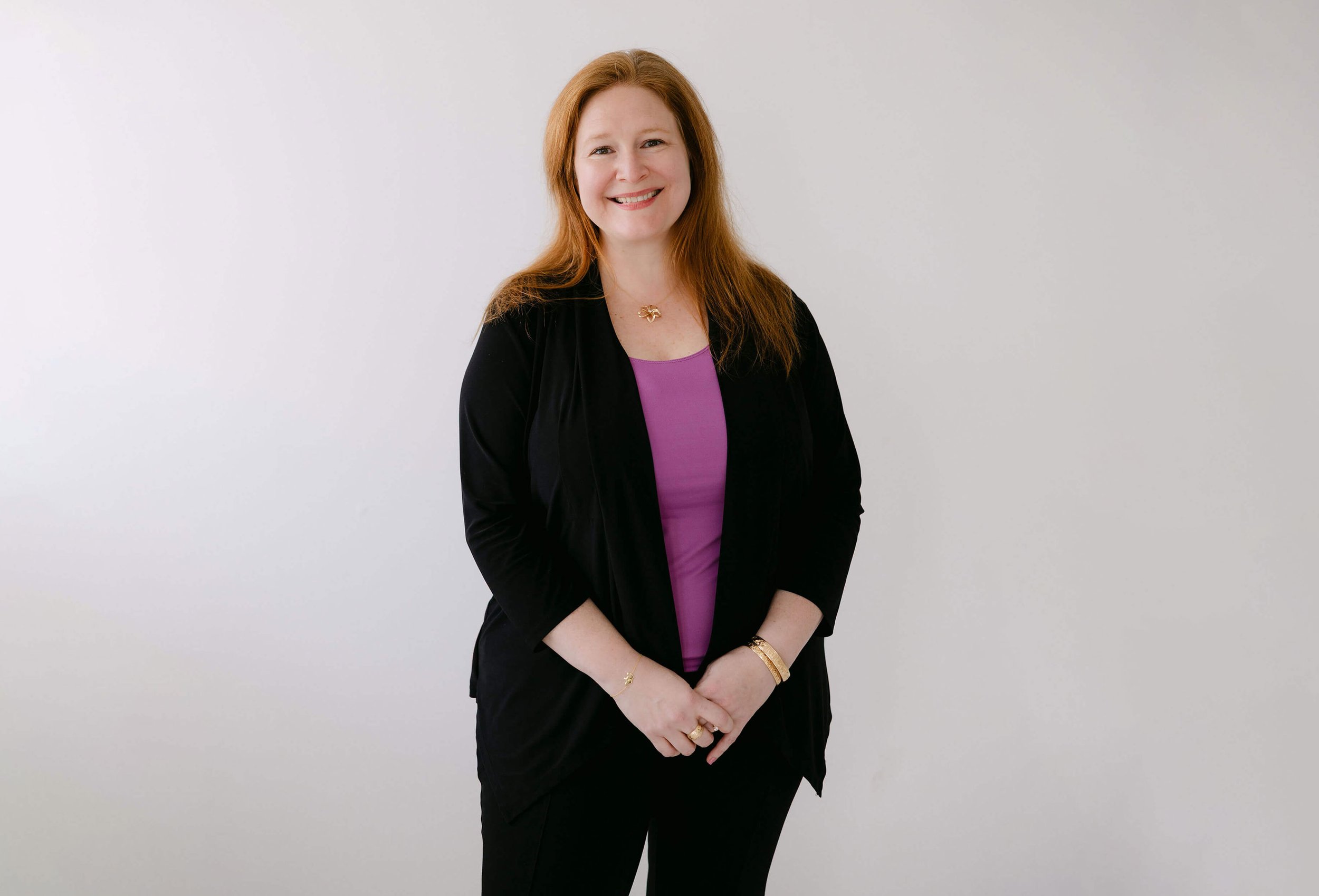 A woman with red hair and fair skin smiling at the camera, dressed in a purple top, black blazer, and black pants, standing against a plain white wall.