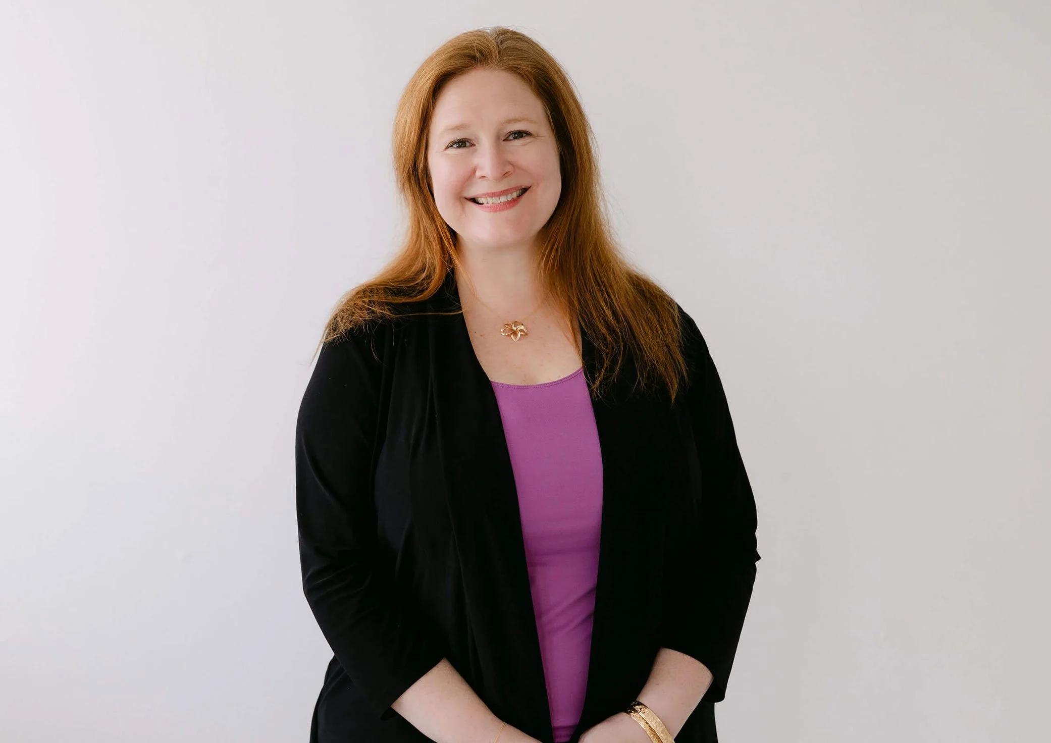 A smiling woman with red hair wearing a purple top and black blazer, standing against a plain white background.
