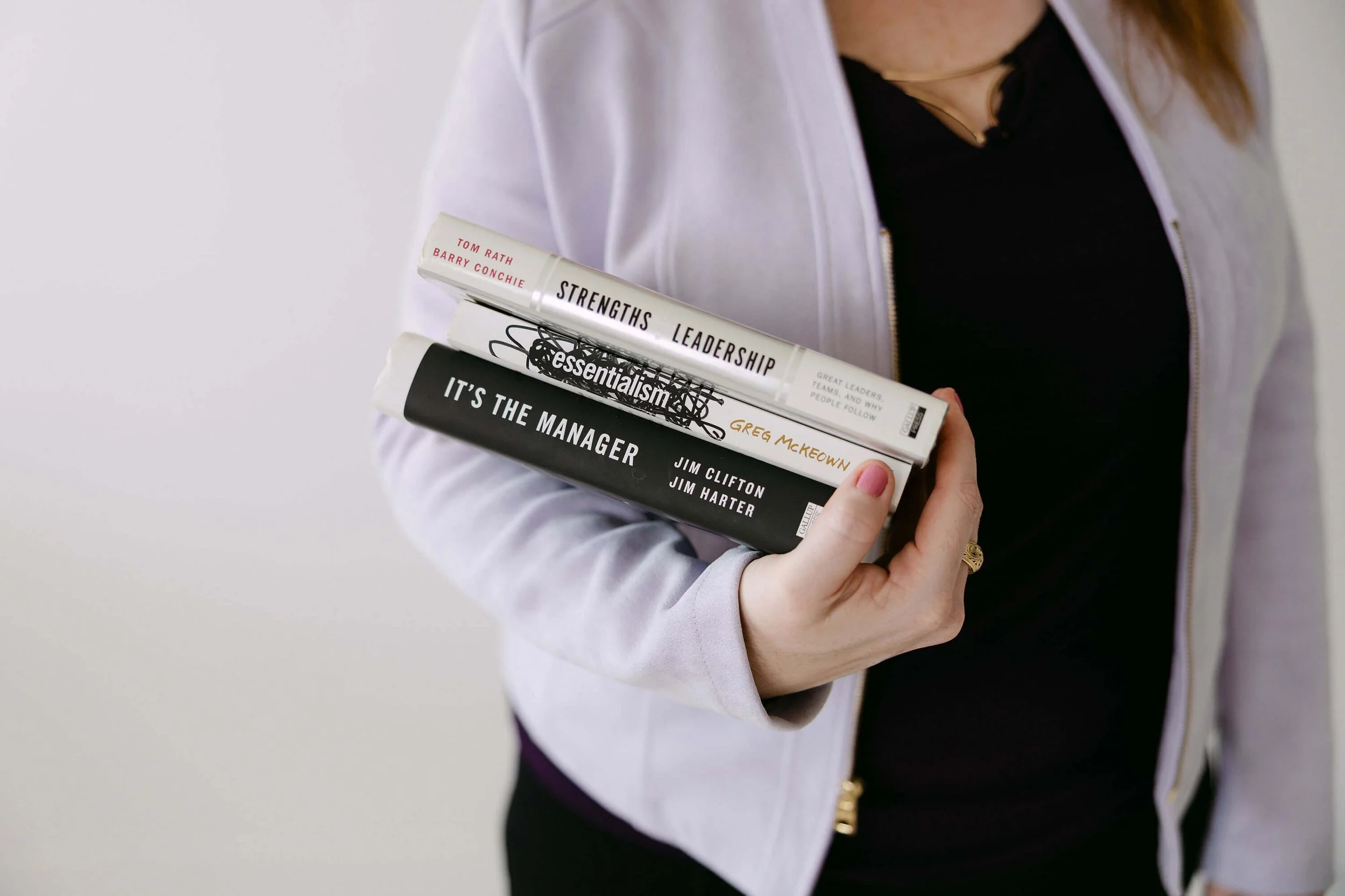 A woman in a white jacket holds a stack of four books on leadership and management.